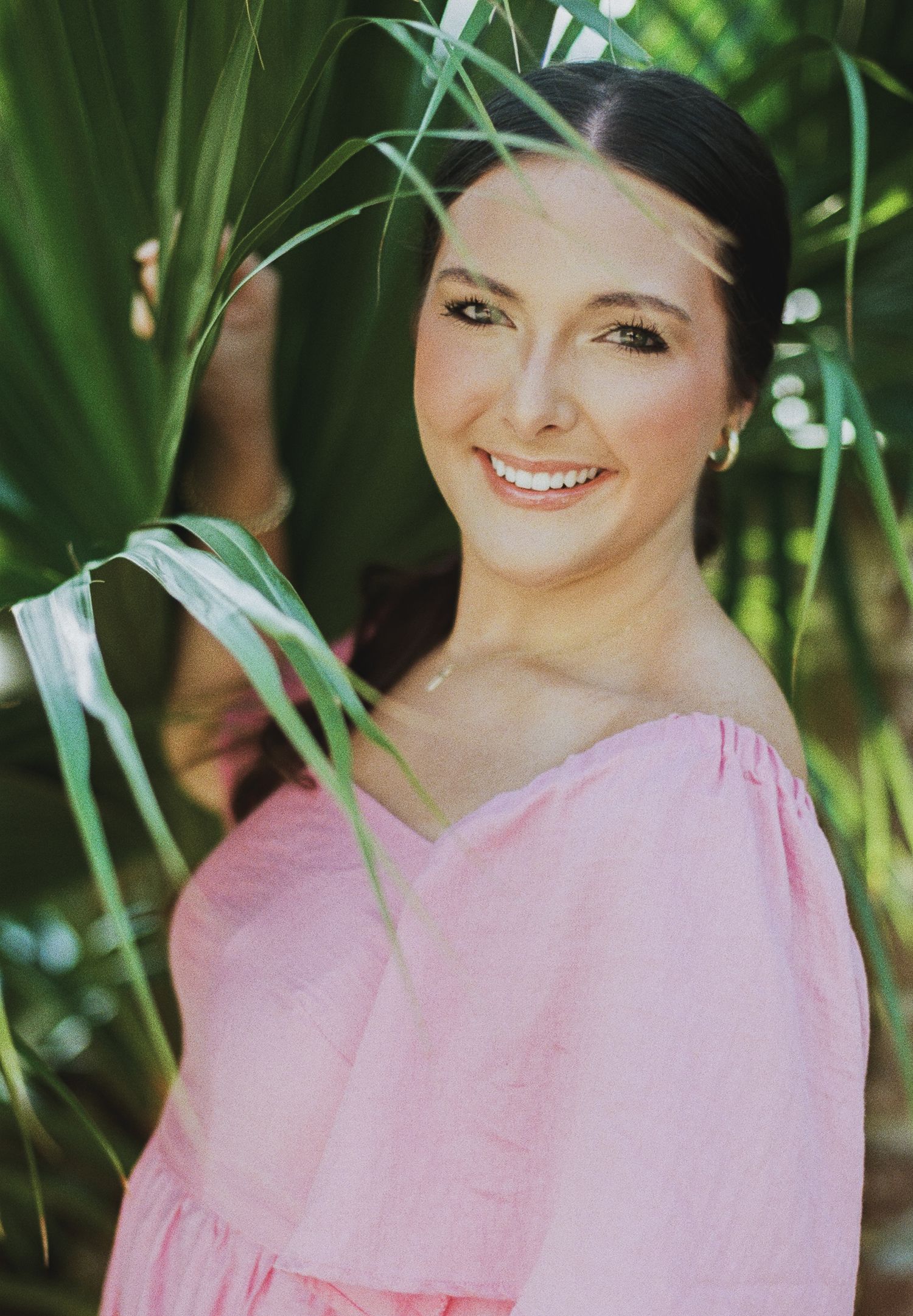 Smiling woman in pink off-shoulder top posing among lush green tropical palm leaves outdoors.
