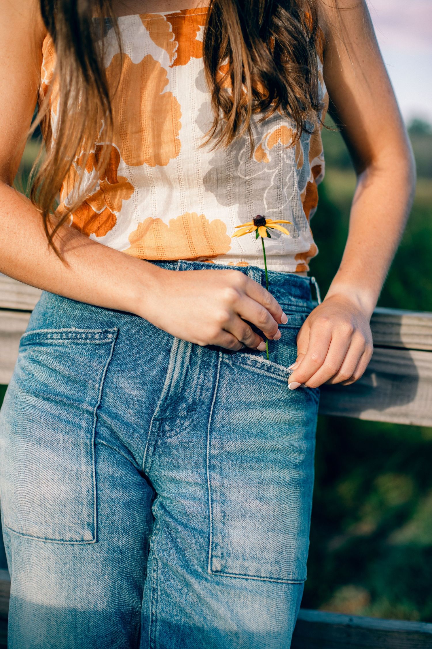 Young woman in floral top and wide-leg jeans holding a yellow wildflower, leaning against a wooden fence outdoors.