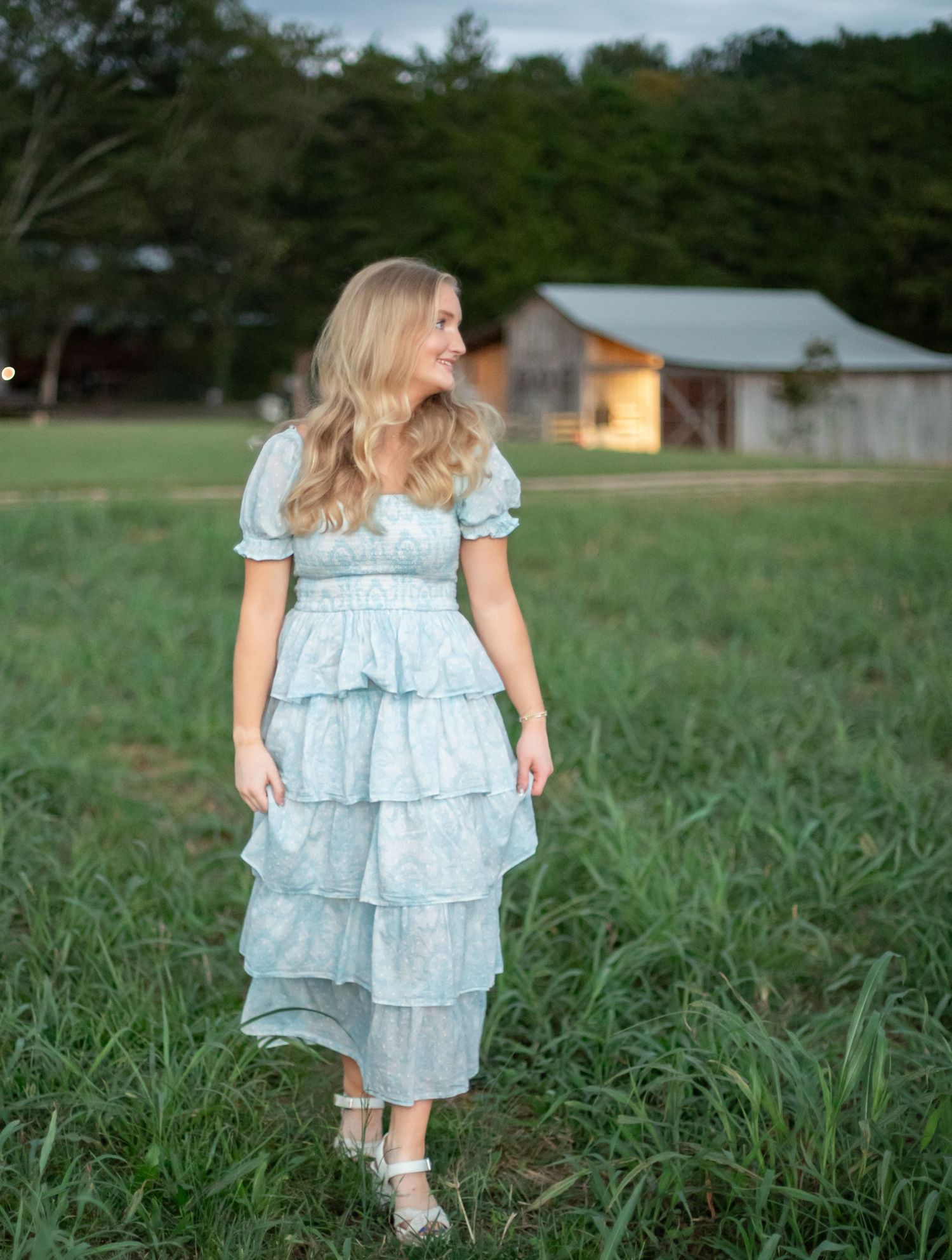 A woman in a light blue tiered ruffle midi dress stands in a green field near a rustic barn at dusk.