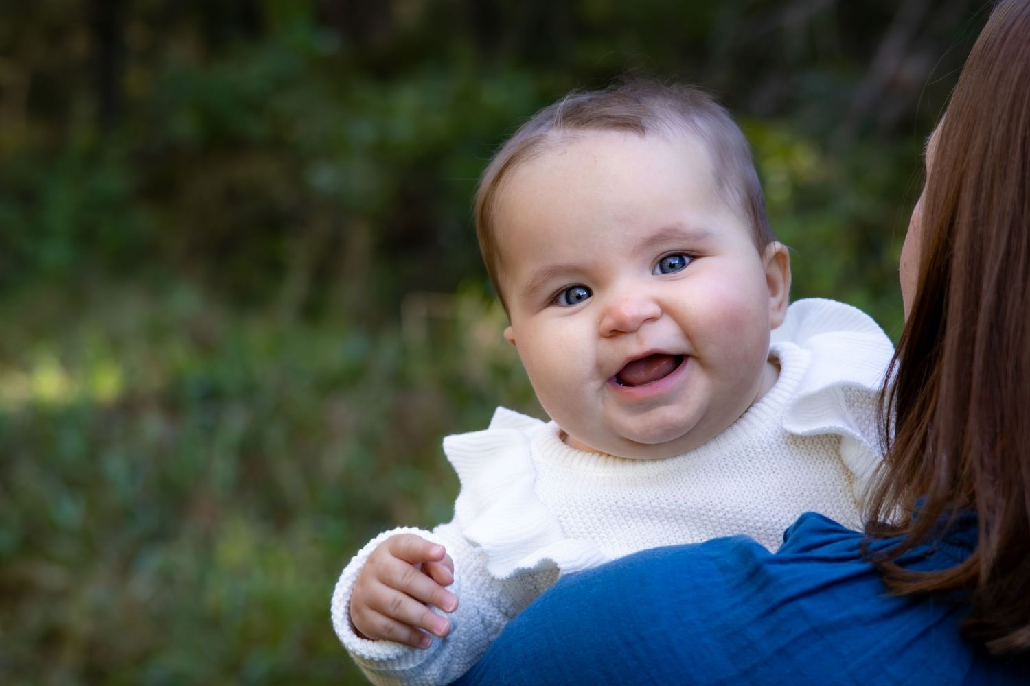 Baby wearing white outfit smiles brightly while being held outdoors.
