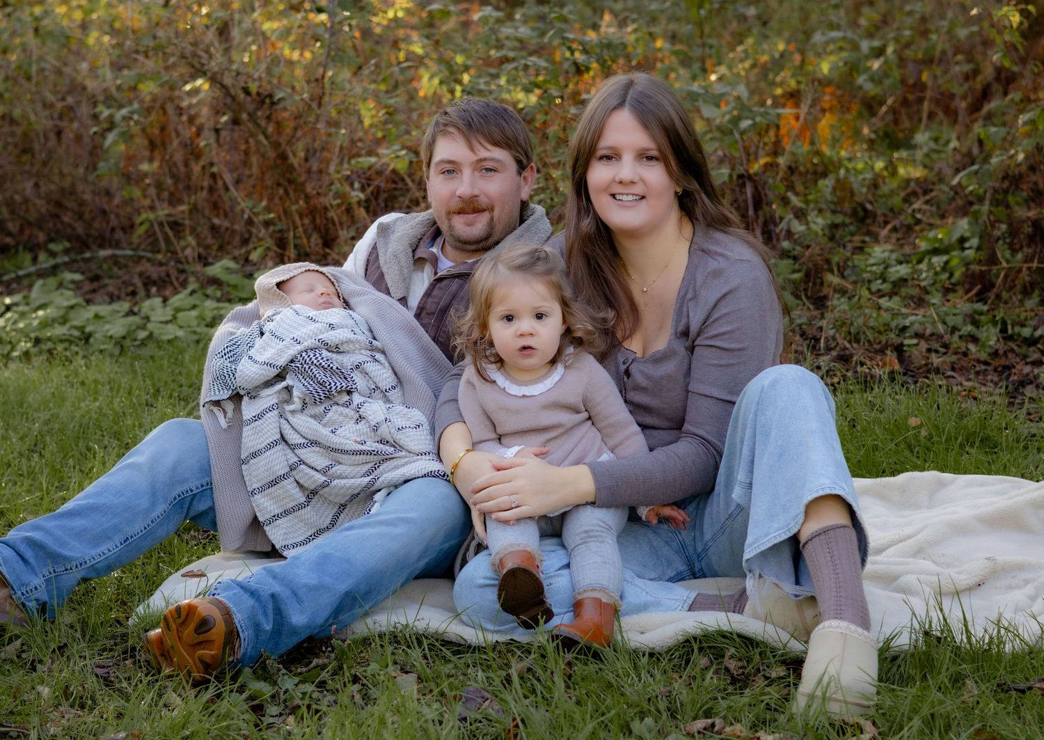 Family lying together on a blanket outdoors during autumn with their dog nearby.