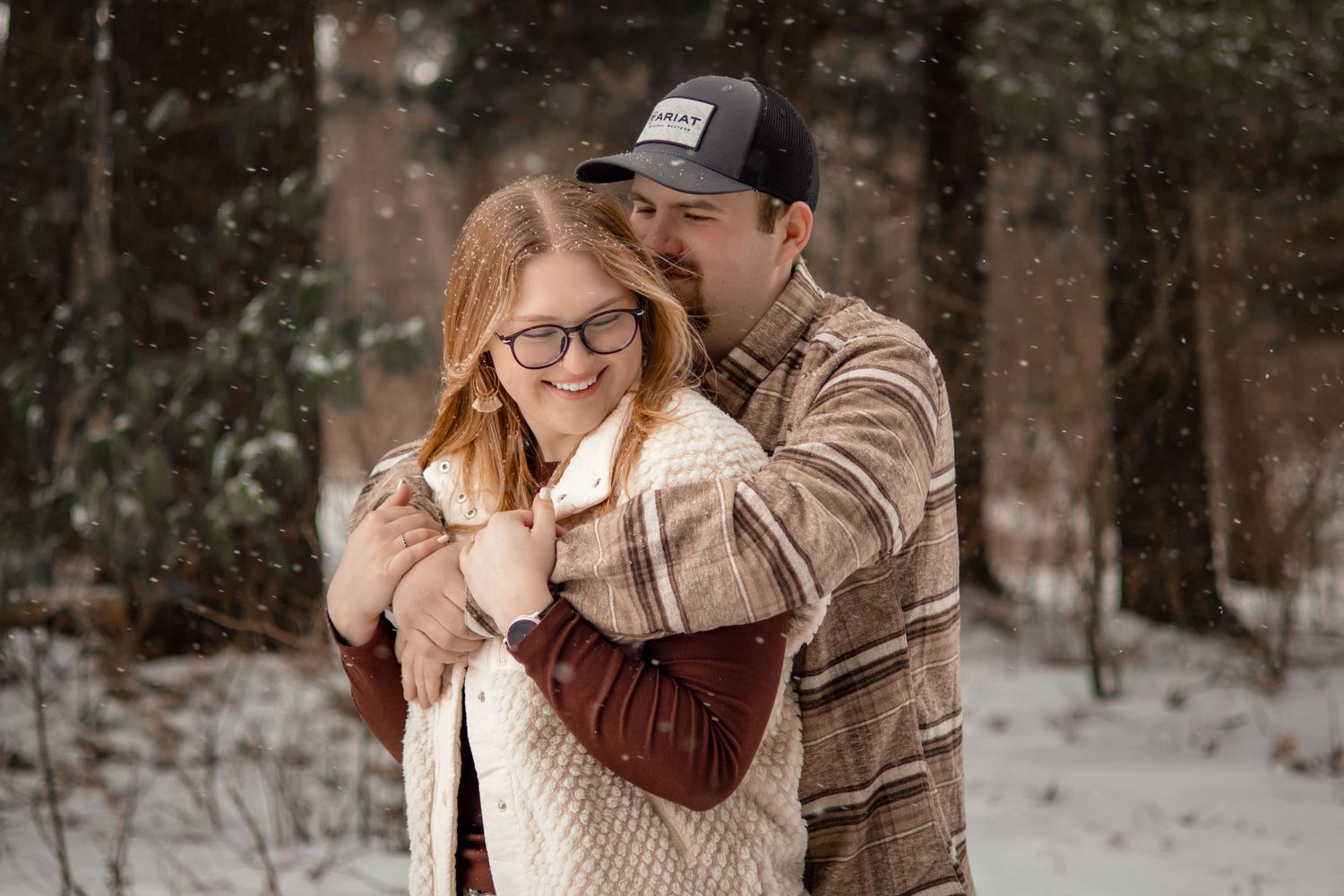 Couple embraces in a romantic winter photoshoot with snow falling around them in a forest setting.