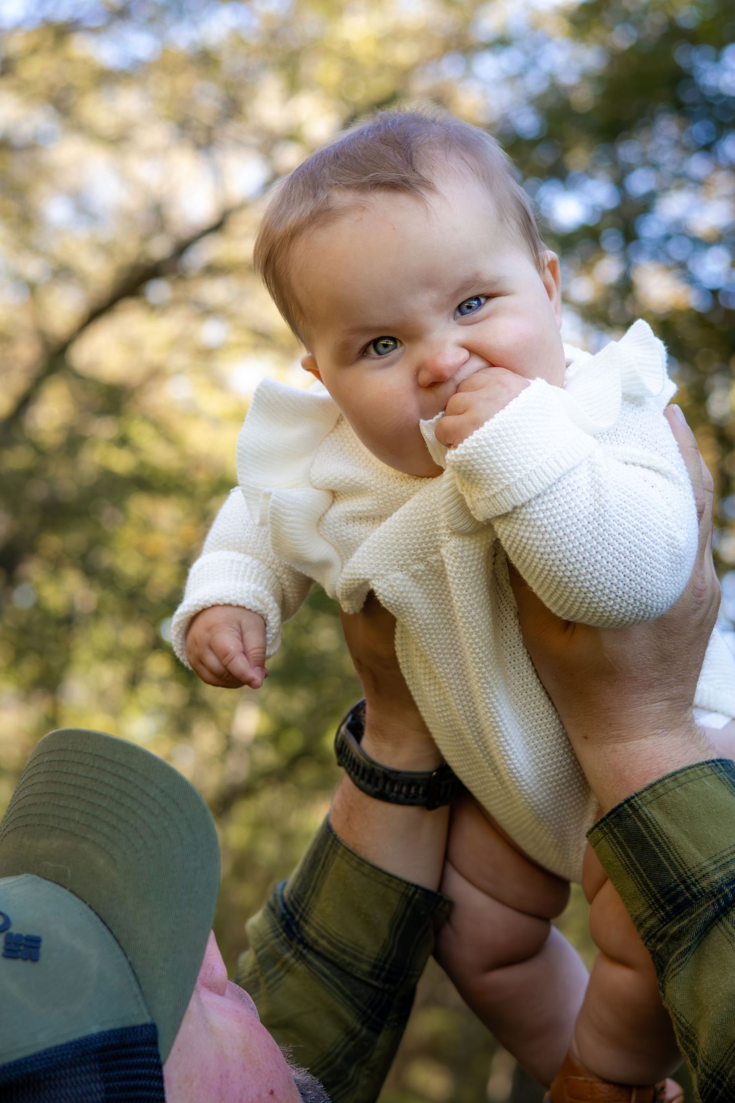 Baby in white sweater being lifted against autumn foliage background.