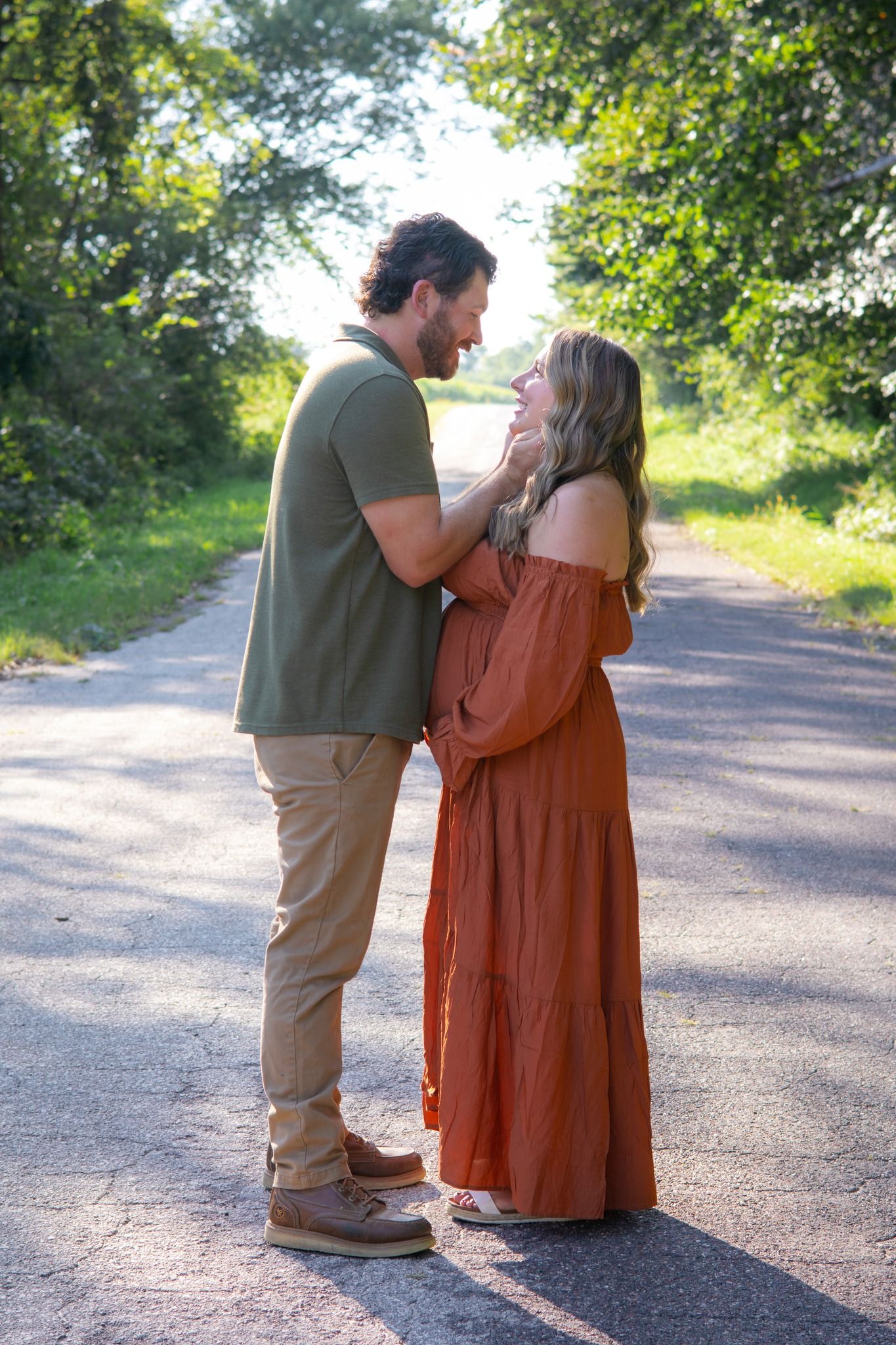 Intimate maternity photo of couple on sunlit path, woman wearing long rust-colored dress.