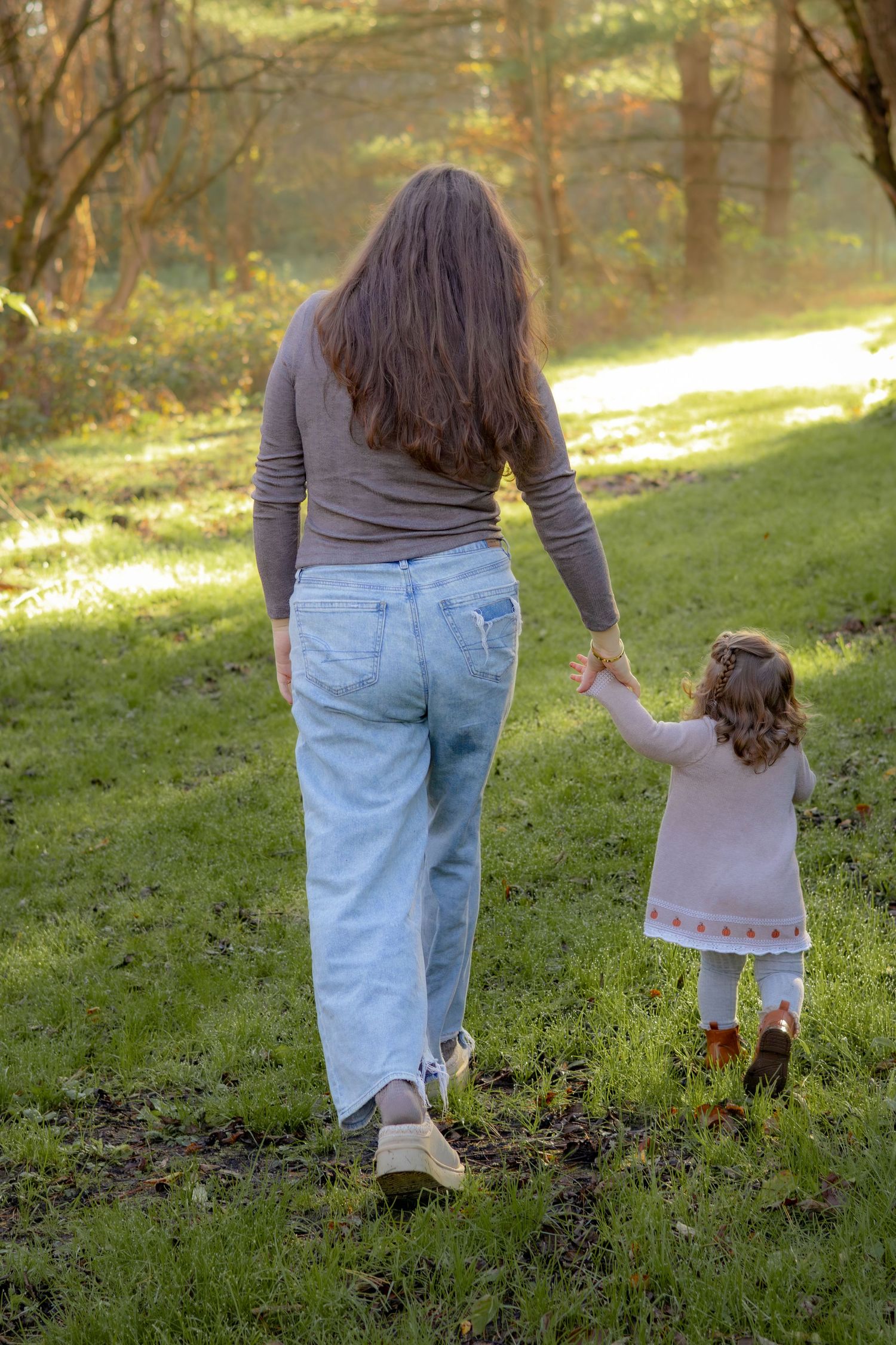 A parent and child walk hand in hand through a sunlit forest path.