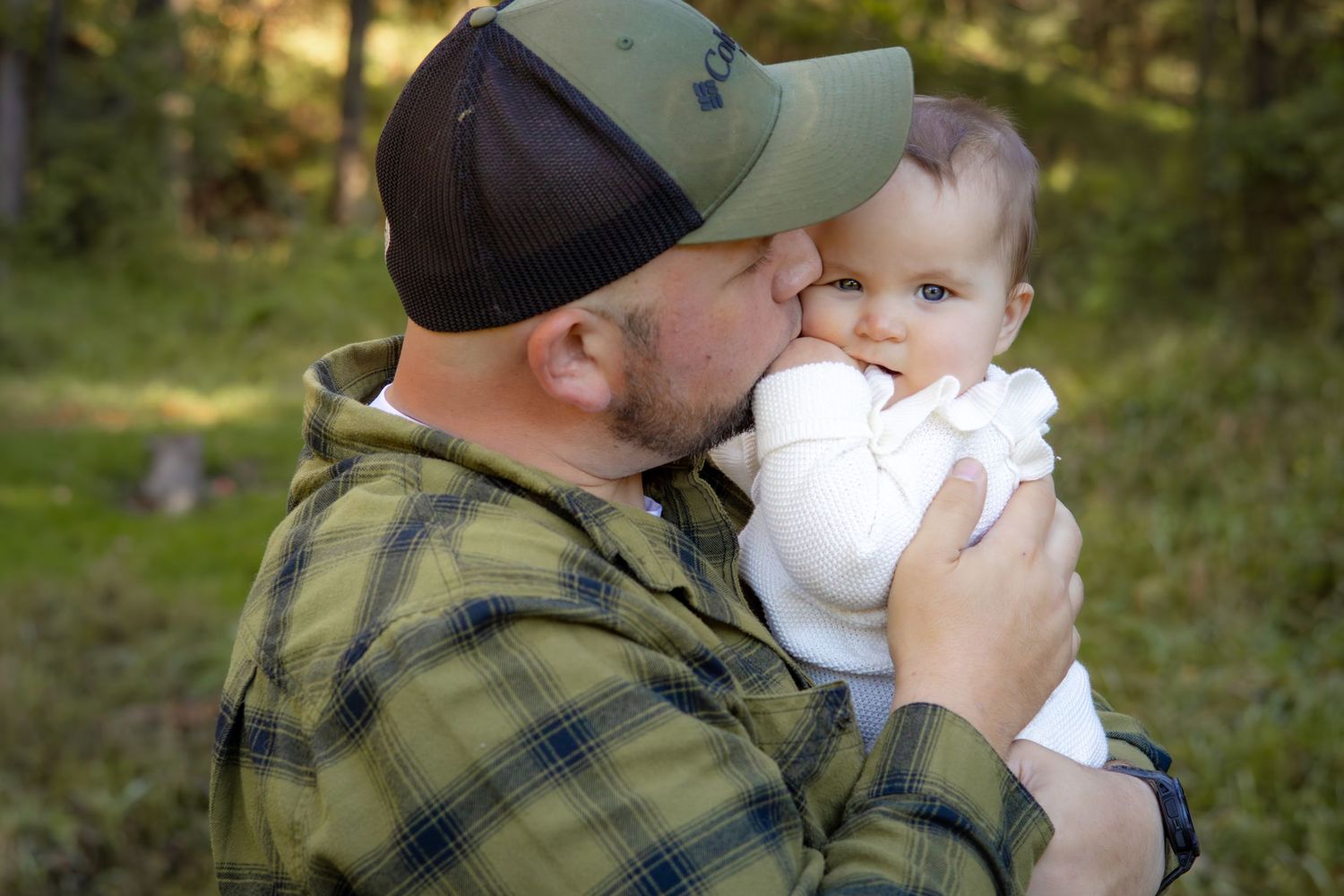 Person wearing plaid and baseball cap holds and kisses small baby outdoors.