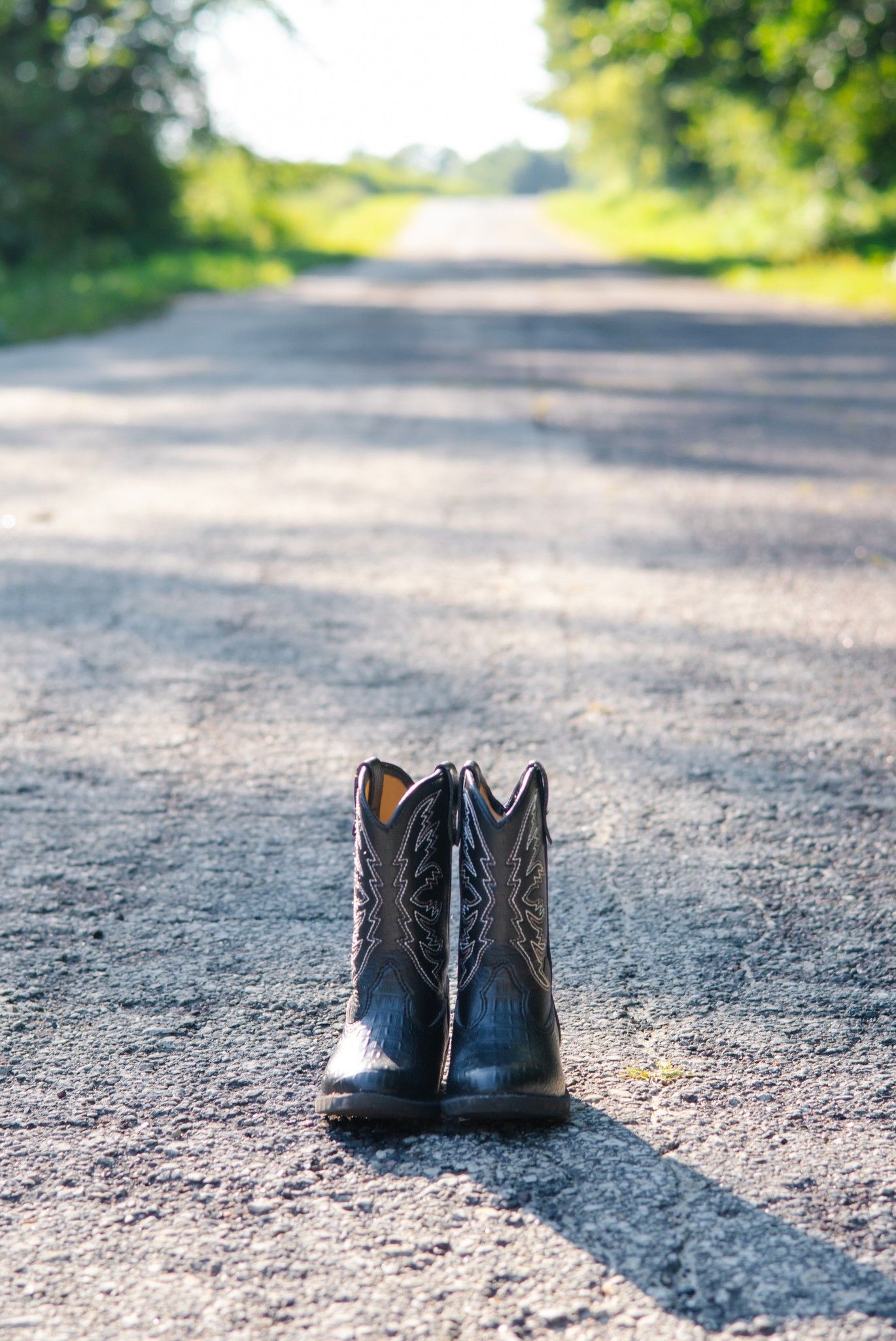 A pair of black cowboy boots standing alone on a dirt road with sunlit path ahead.