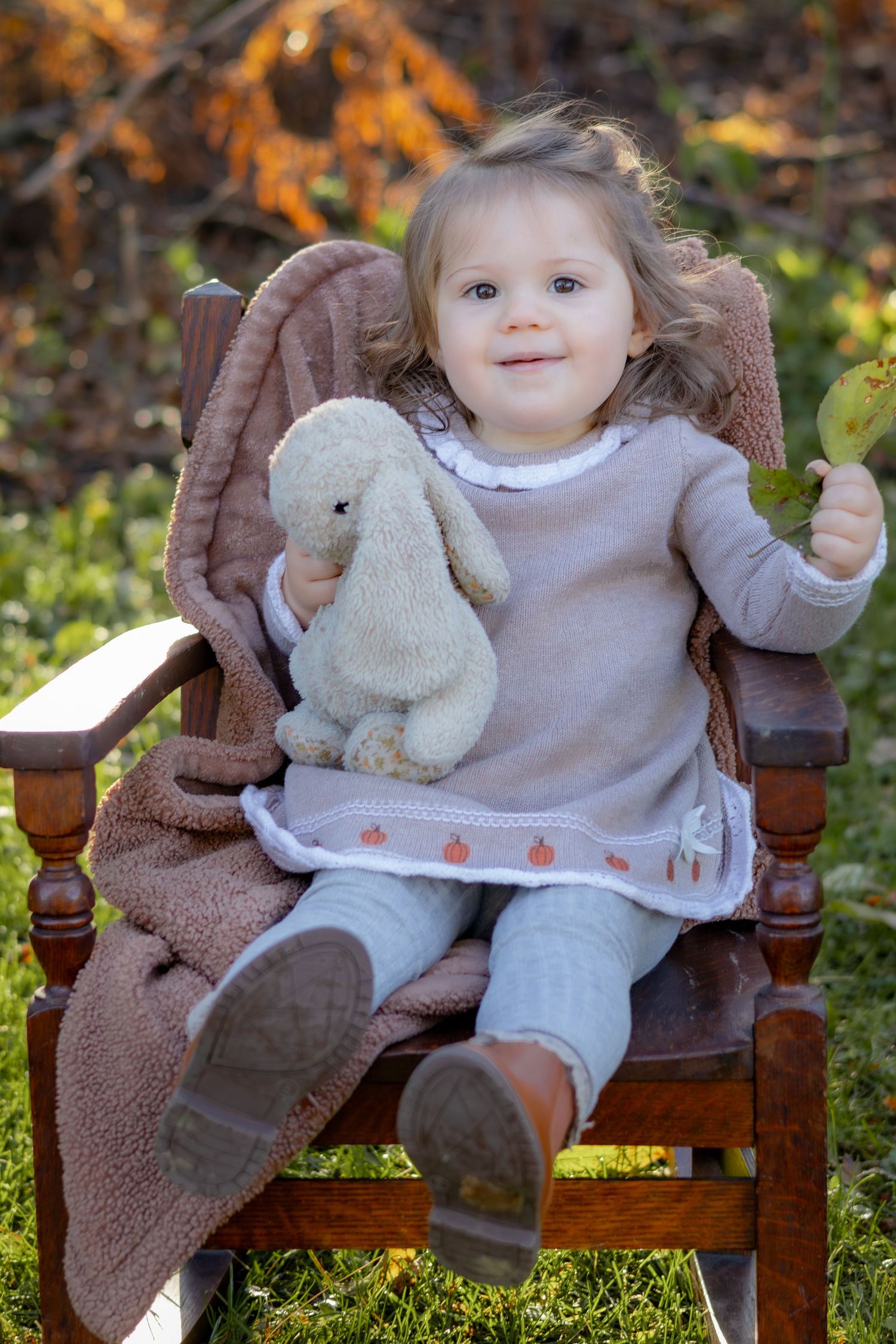 Toddler sitting in wooden chair holding stuffed bunny in autumn setting.