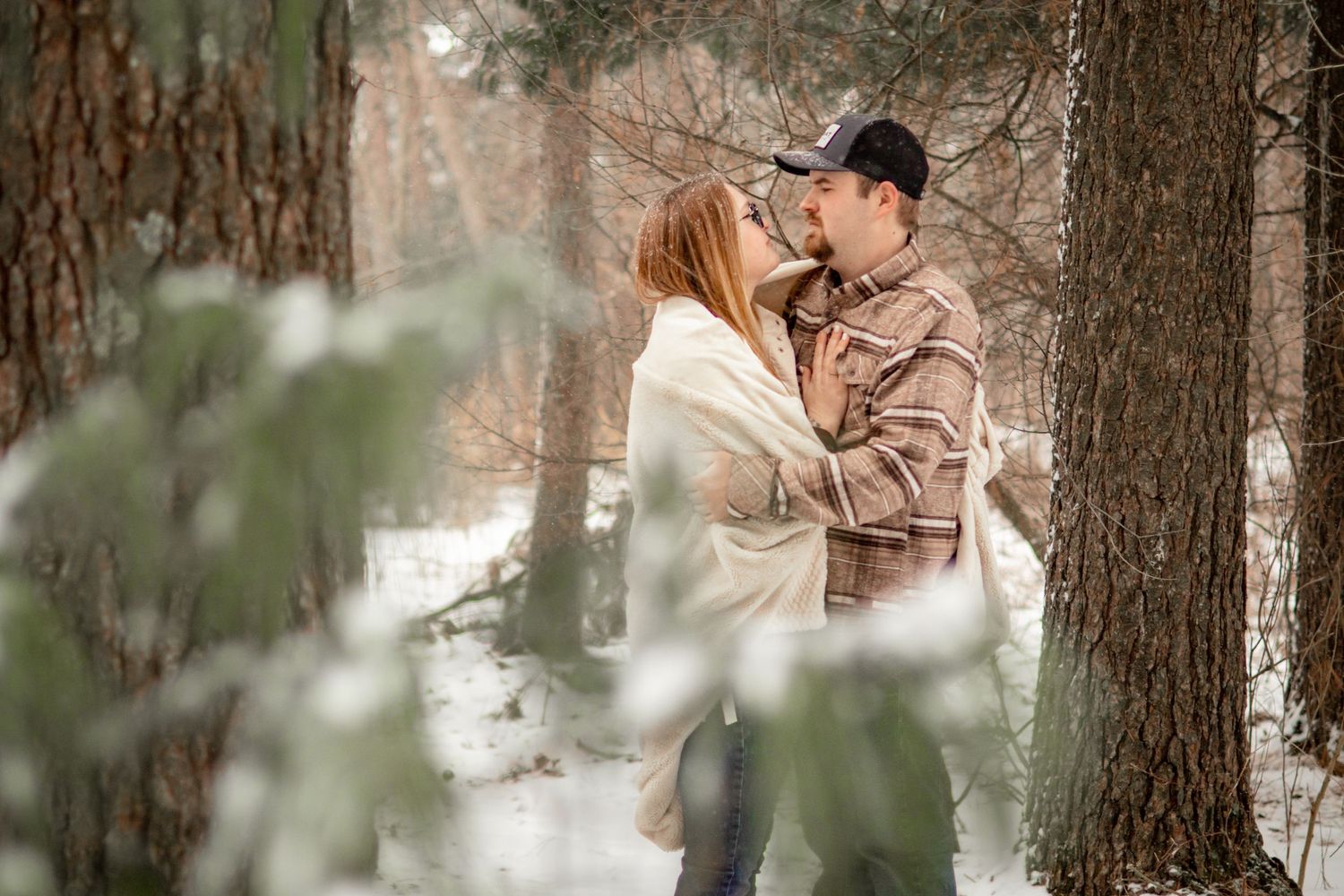 Couple embracing in snowy winter forest scene framed by evergreen branches.