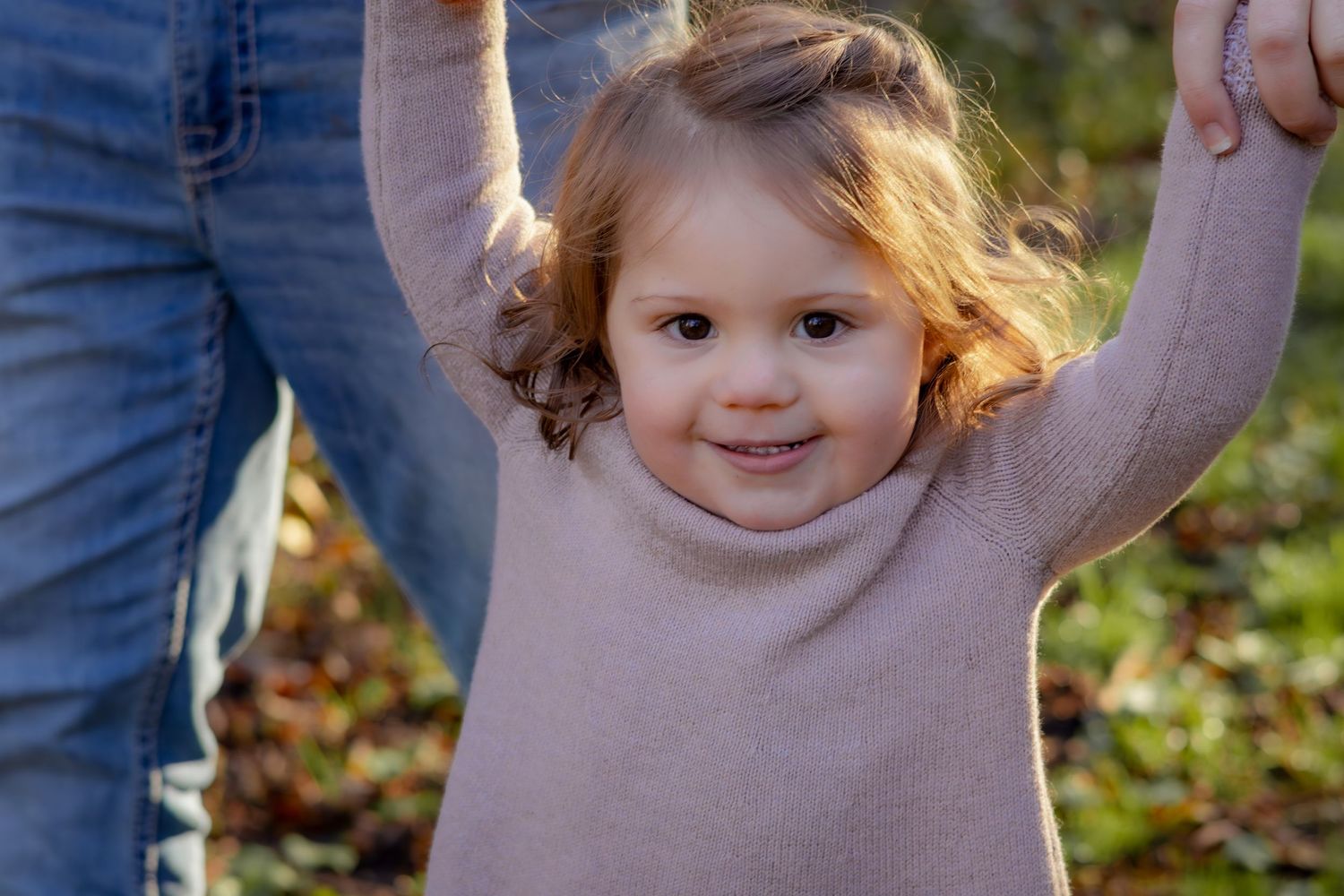 A joyful toddler raises their arms while standing outdoors in autumn.
