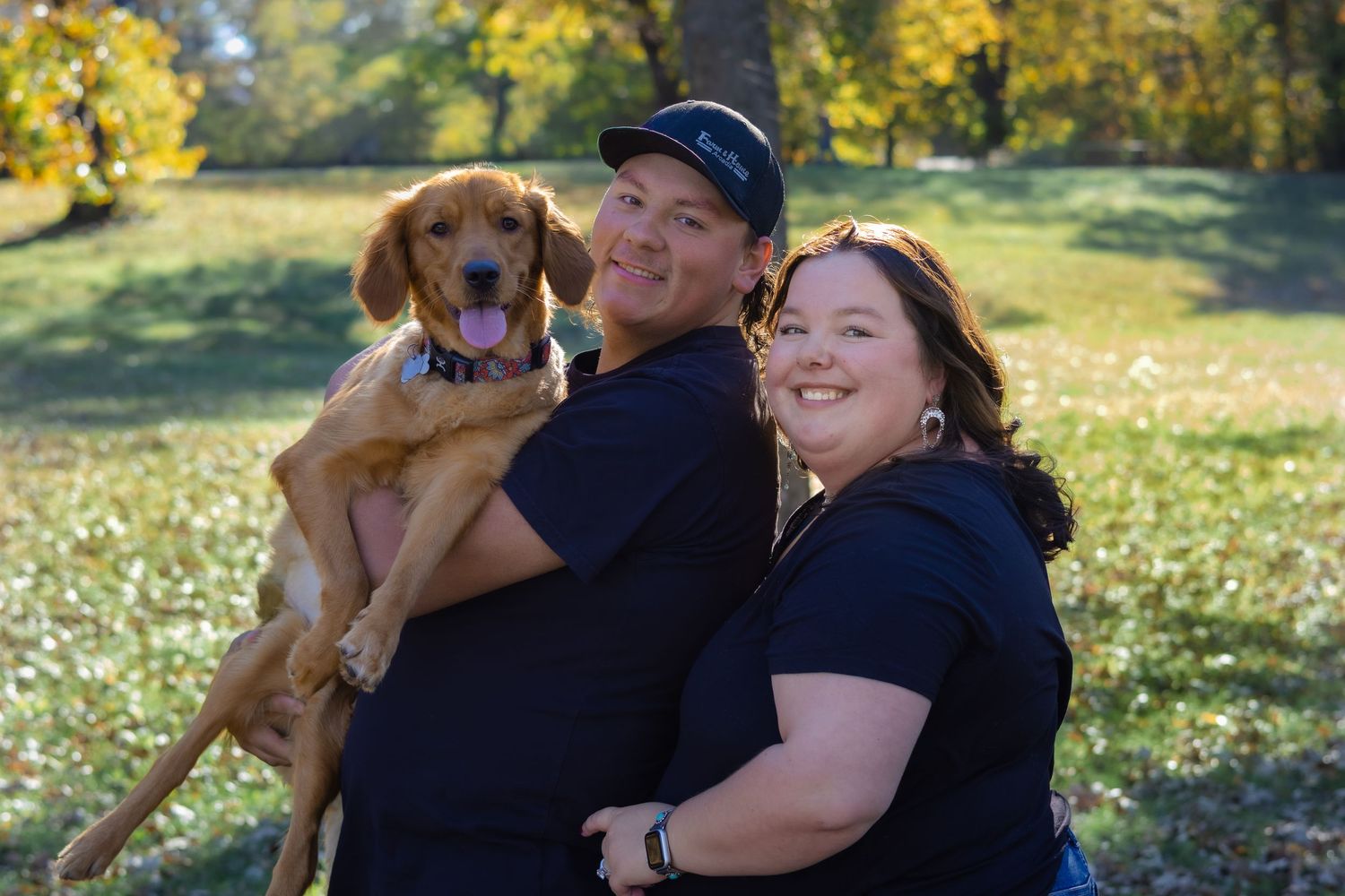 Couple posing with their golden retriever dog in a sunny meadow.