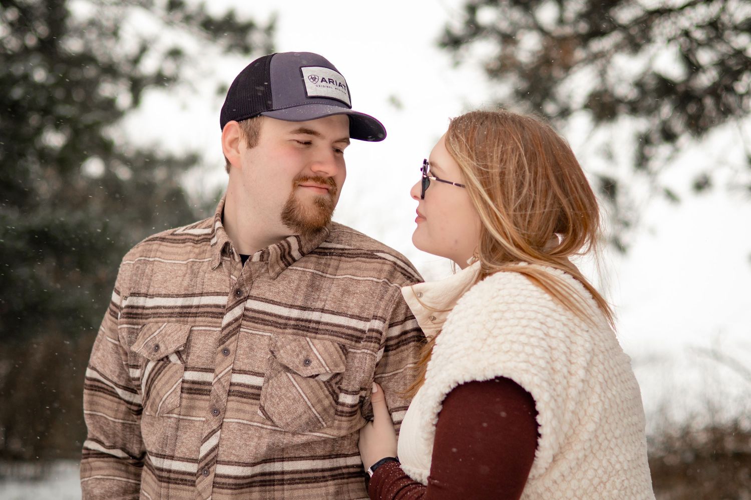 A couple embraces in a snowy winter forest setting wearing casual clothing.