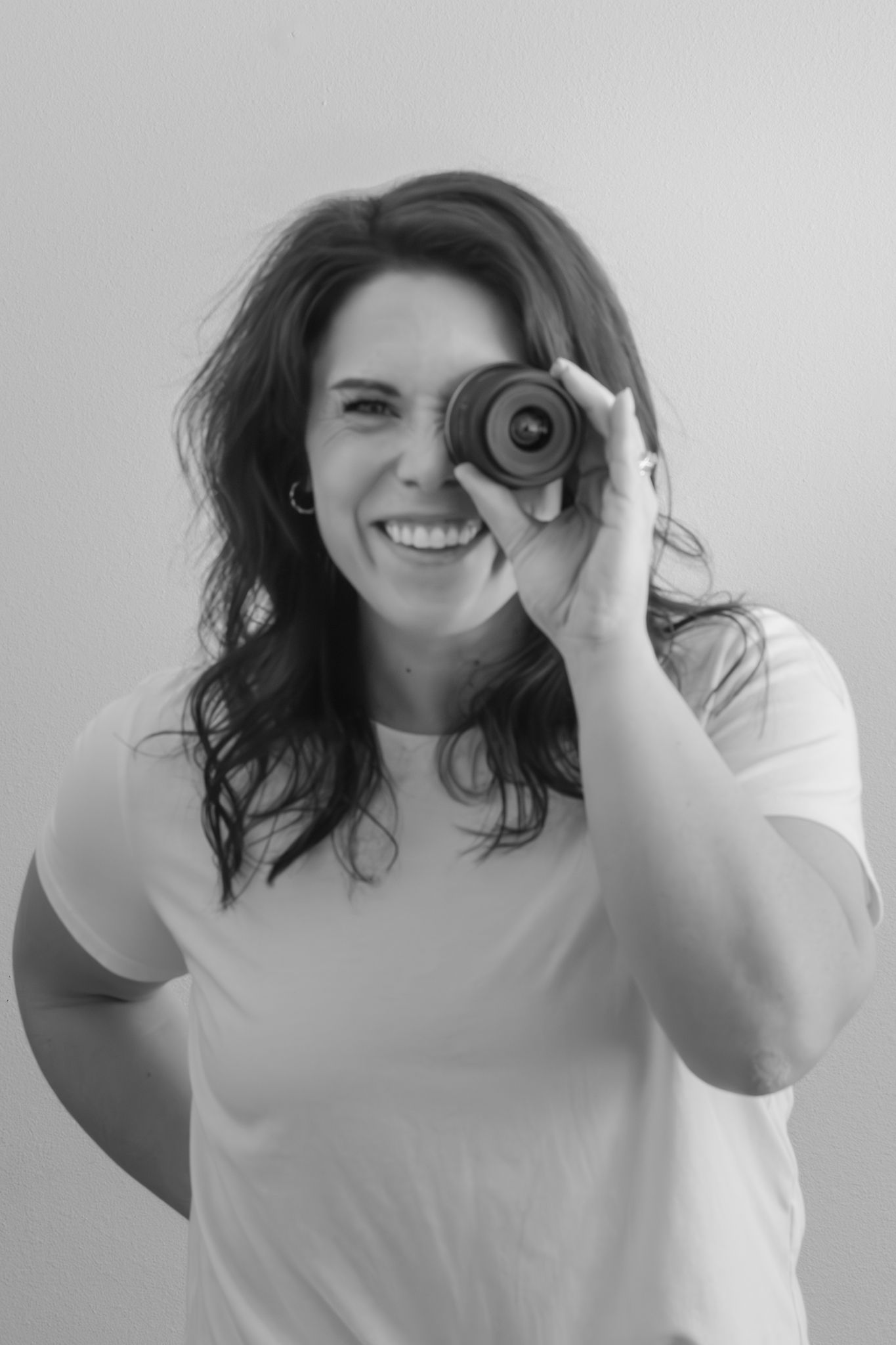 Black and white portrait of a photographer looking through a camera lens with a bright smile.