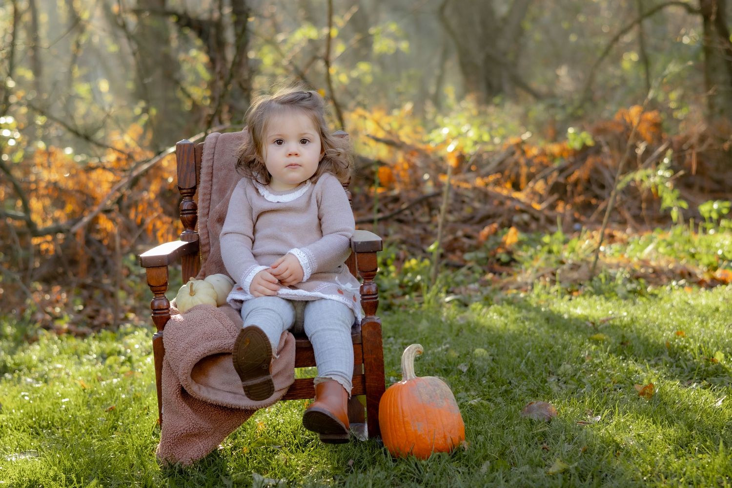 A toddler sits in a brown chair next to a pumpkin in an autumn garden setting.