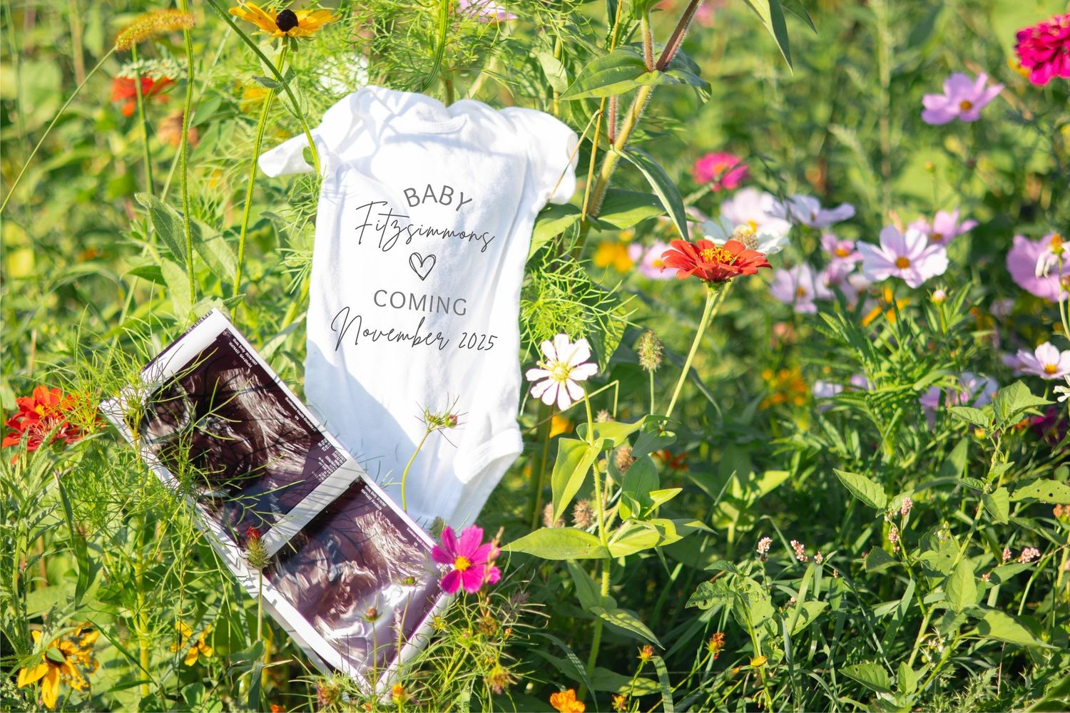 Baby onesie and ultrasound photo displayed among colorful wildflowers in a garden setting.