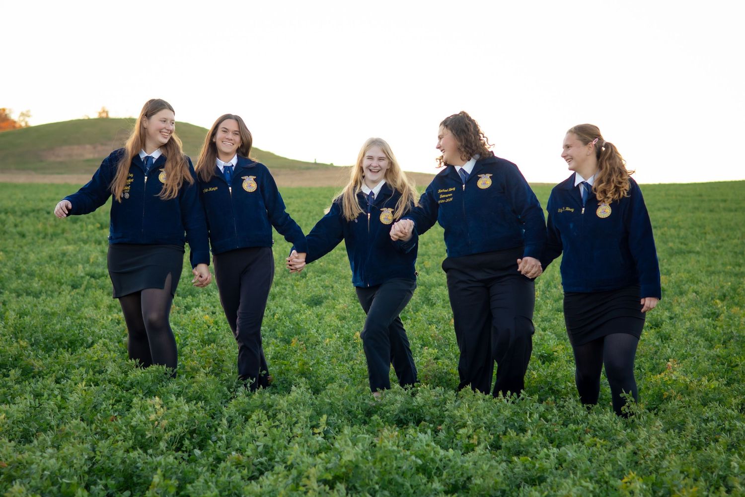 Group of people in dark clothing walking through green field holding hands.