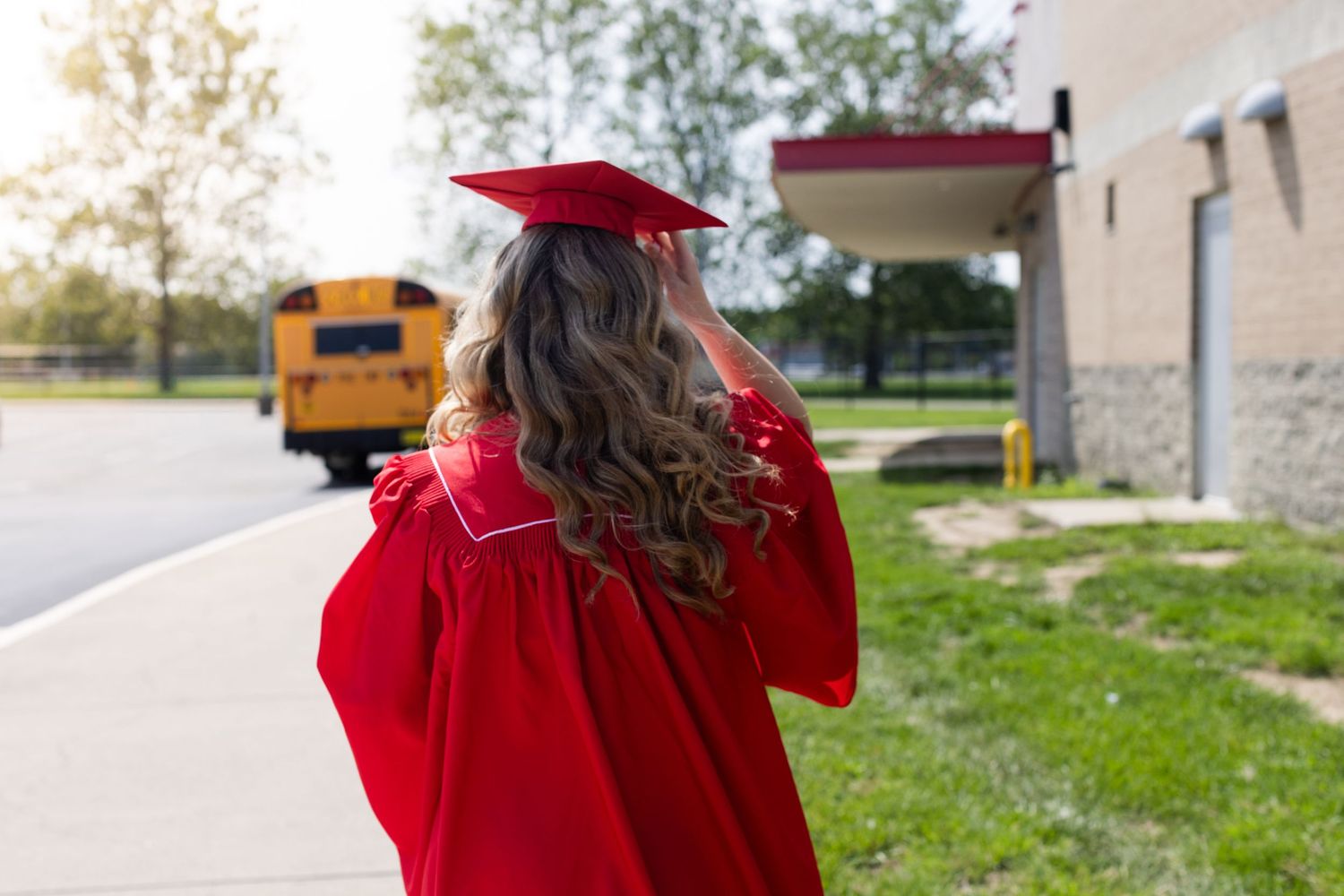 A female graduate in red cap and gown stands outside a school building with a yellow school bus in the background.