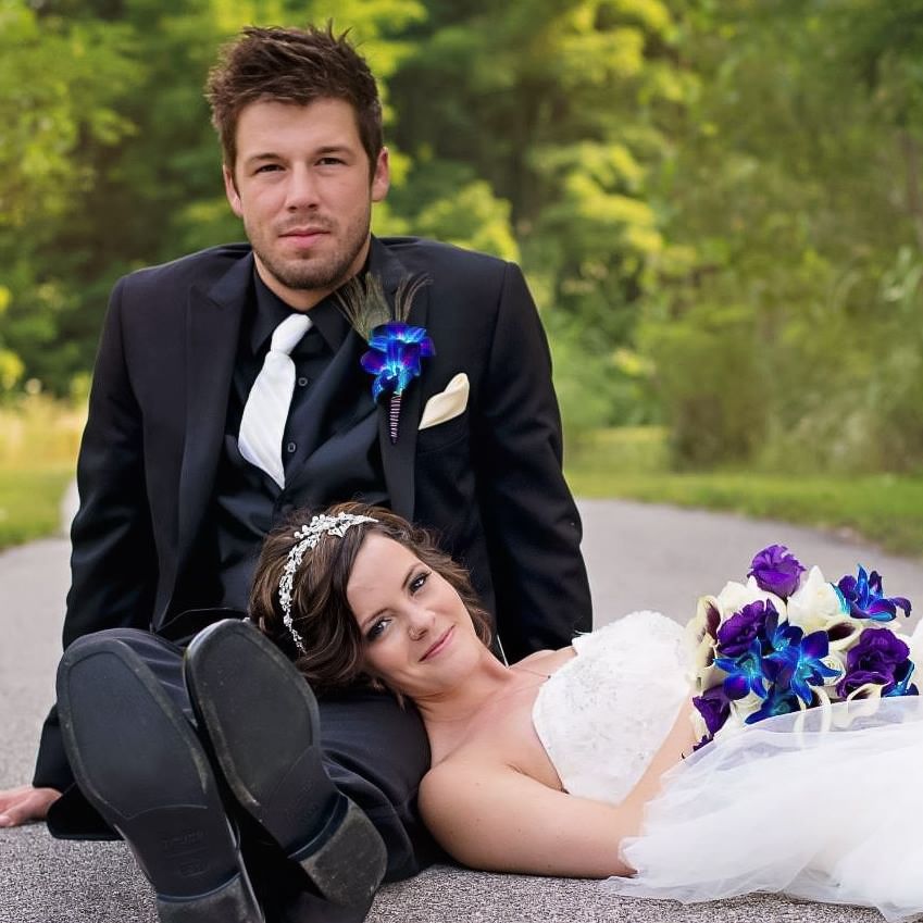 Wedding couple posing on road, bride holding purple bouquet, groom in black suit.