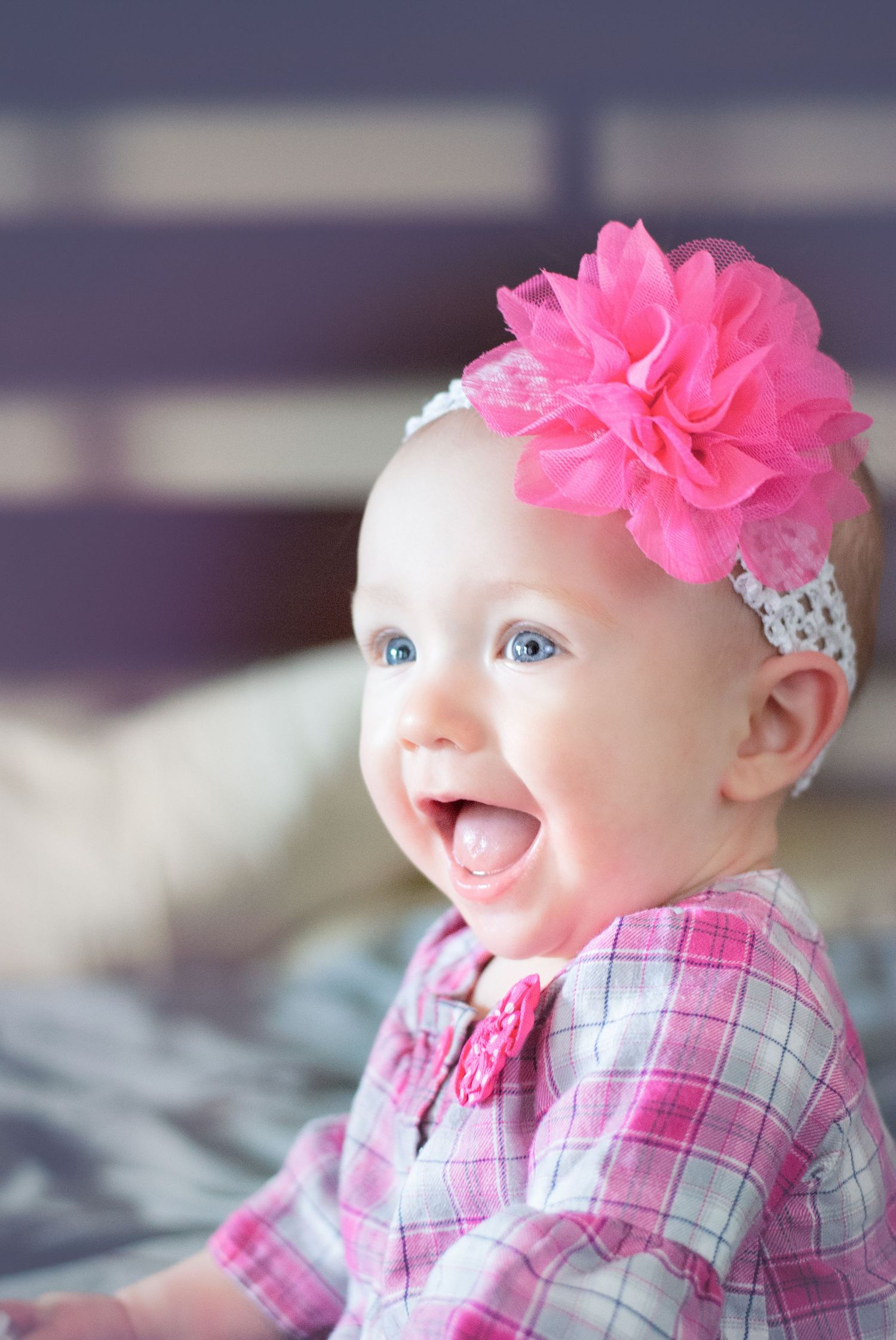 Smiling baby girl with blue eyes wearing a pink floral headband and pink plaid outfit.
