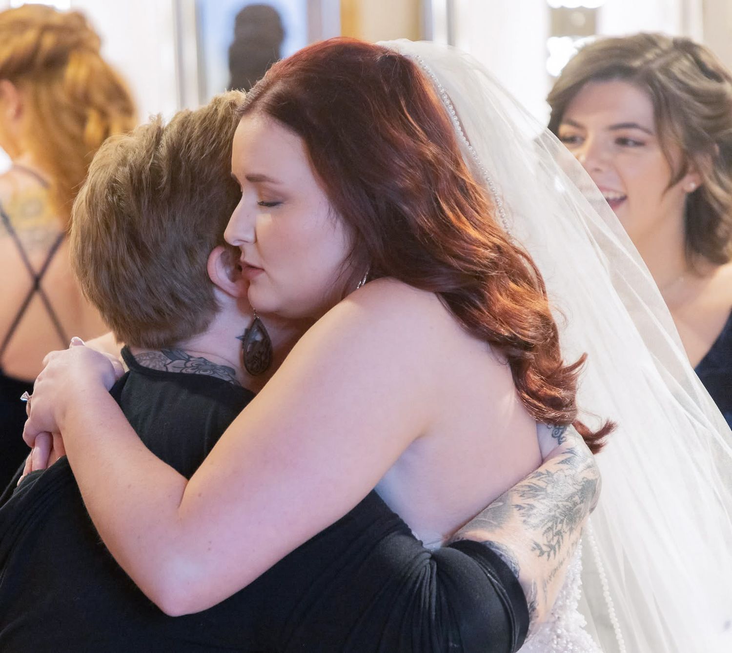 Emotional bride in veil hugging woman in black dress with bridesmaid smiling in background.