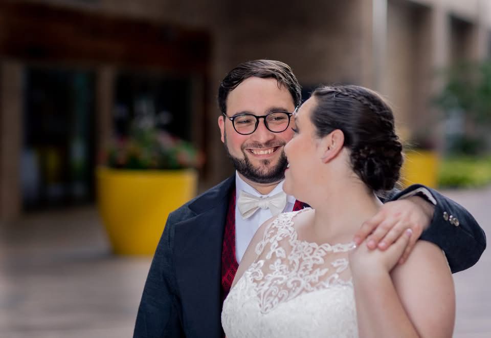Groom in navy suit with glasses smiles as bride in lace wedding dress embraces him outdoors.