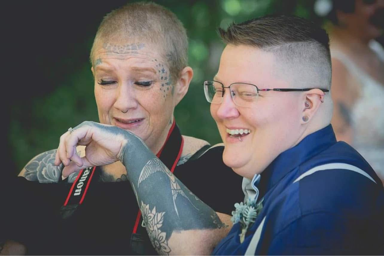 Two brides laughing joyfully during outdoor wedding ceremony, one wiping tears with tattooed arm.