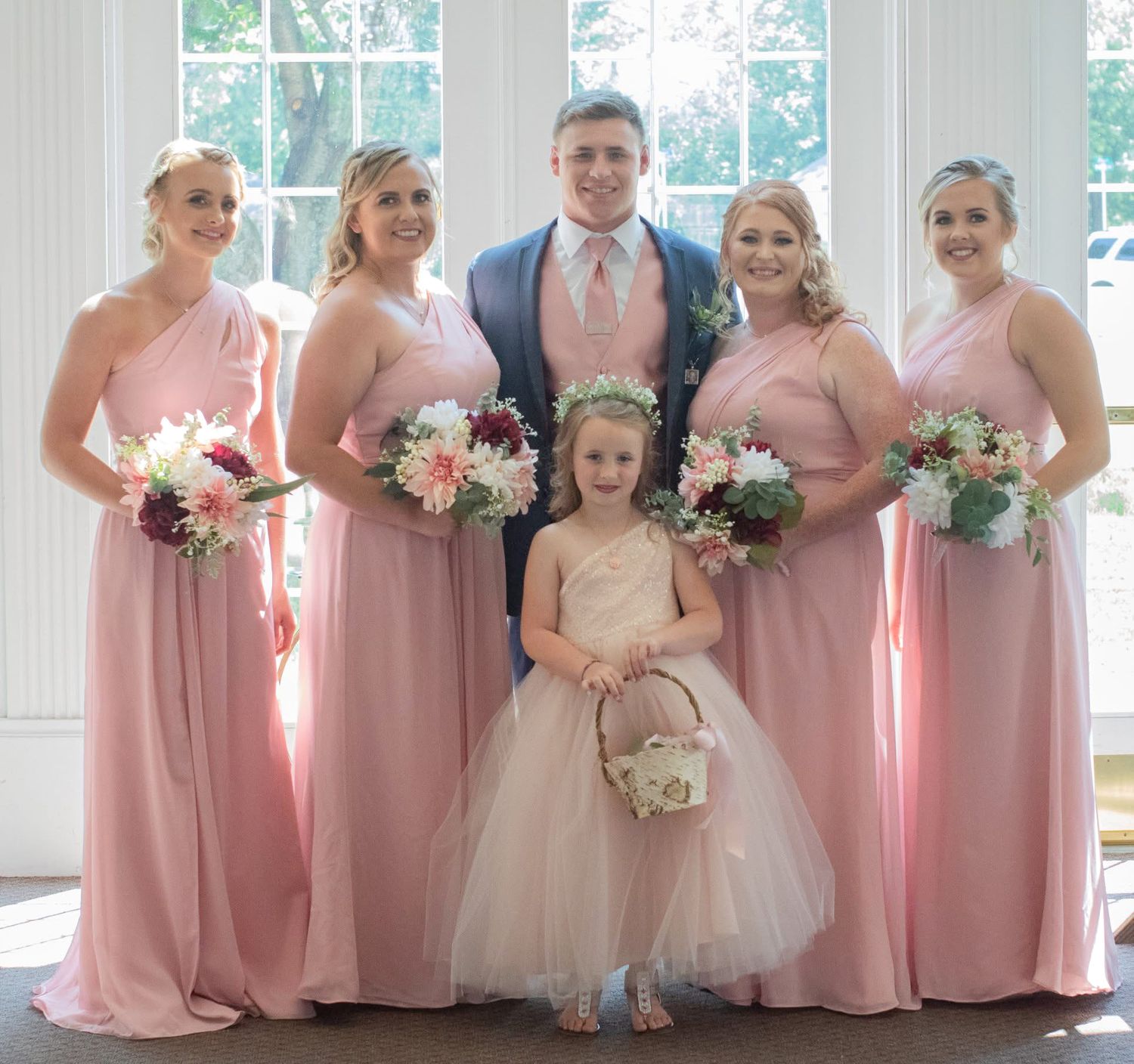 A groom in a blue suit poses with four pink-gowned bridesmaids and a flower girl.