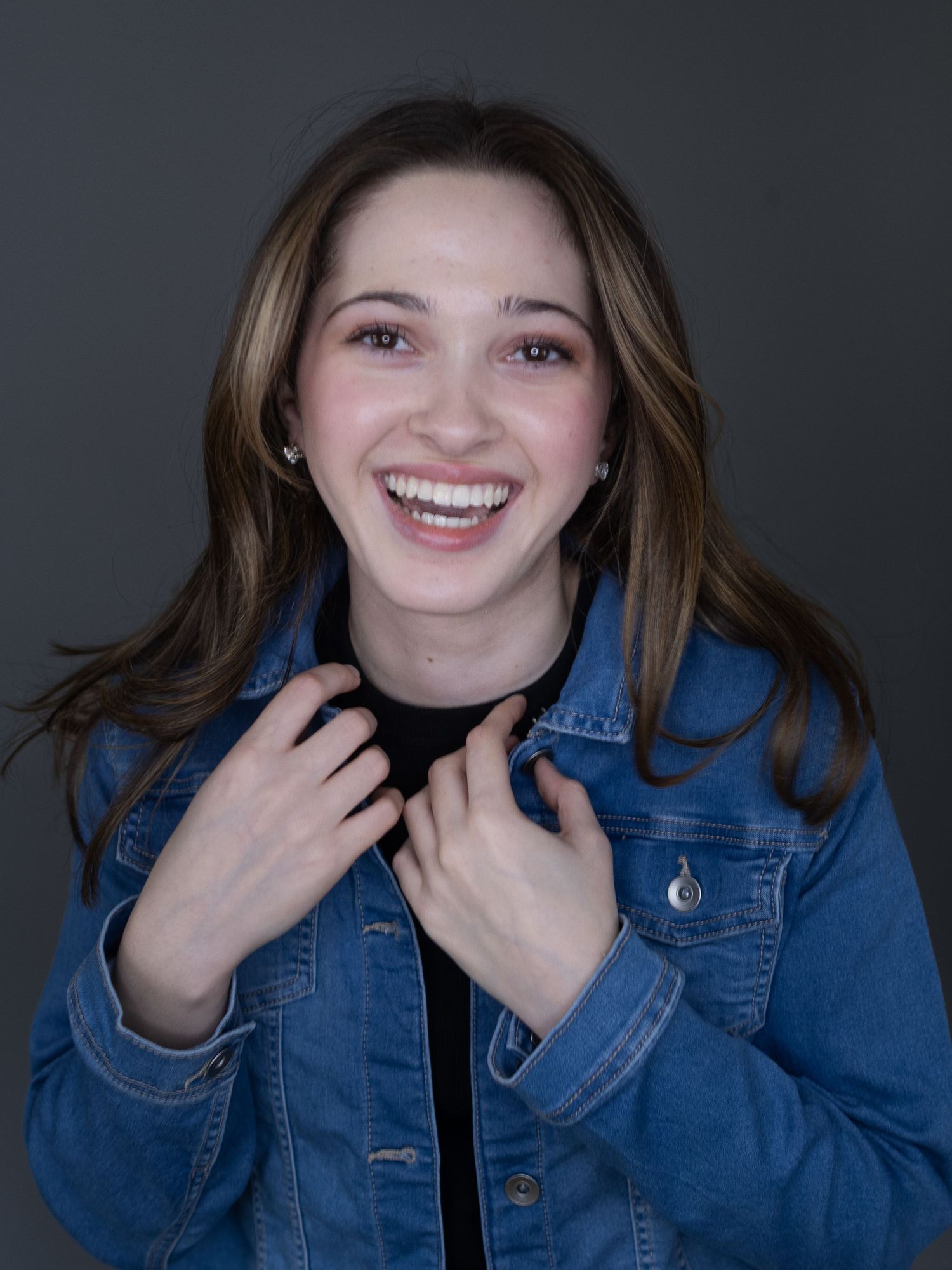Smiling young woman with brown hair wearing a denim jacket, posing against a gray background.