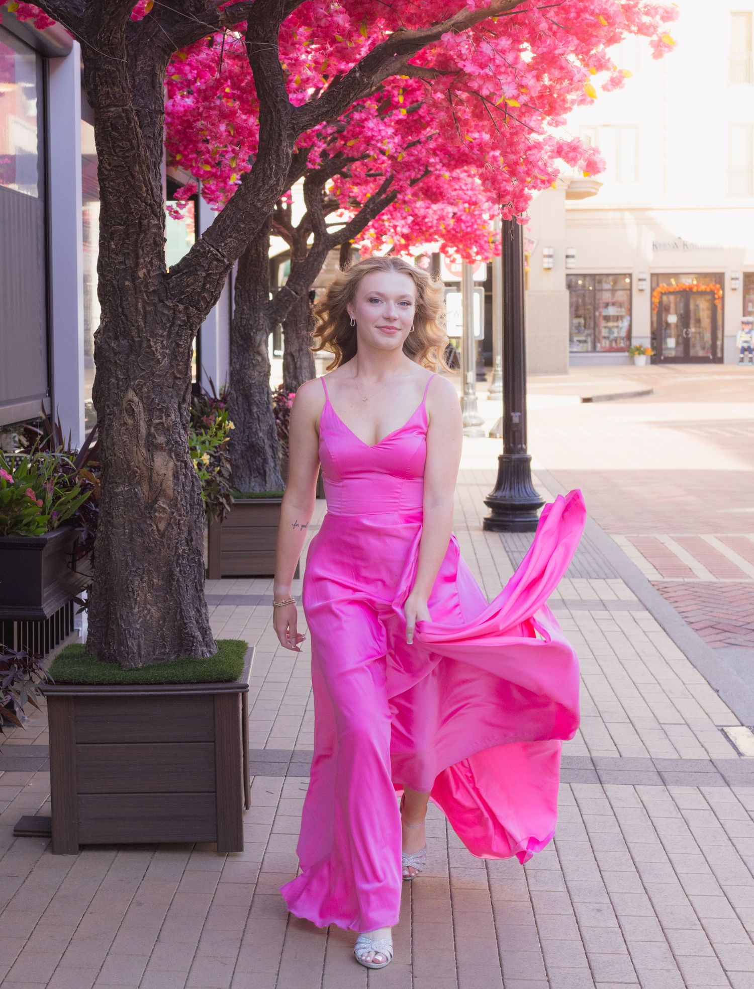Woman in flowing pink satin jumpsuit walking on urban sidewalk beneath vibrant pink flowering tree.
