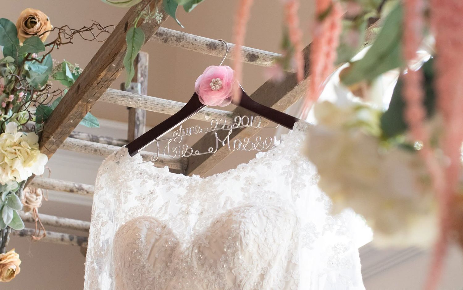 A lace wedding dress hangs on a personalized wooden hanger surrounded by floral decor.