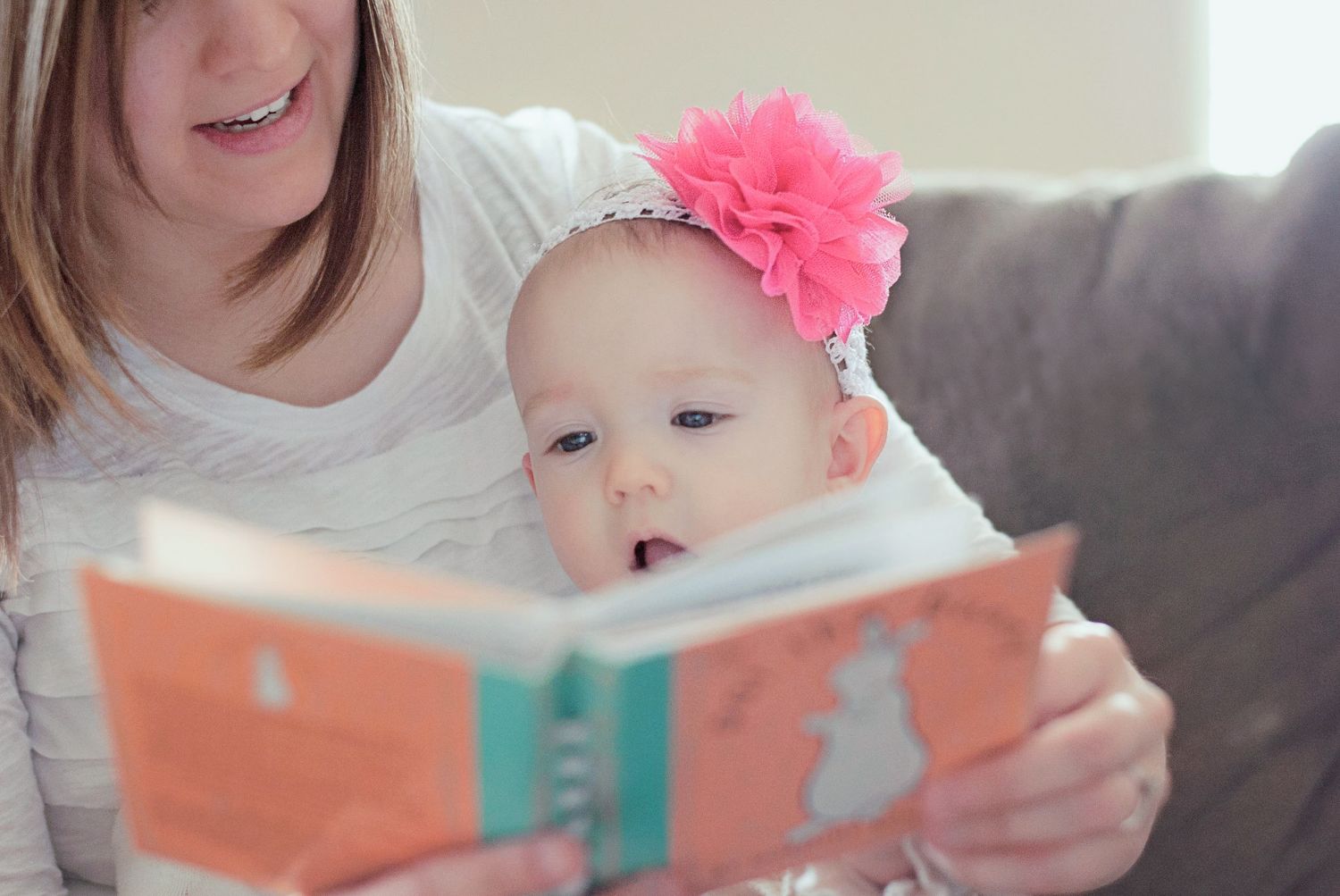 Young girl with braces reads colorful book to smiling baby with pink flower headband on sofa.