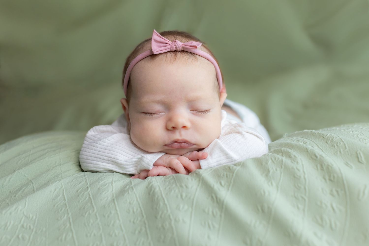 Peaceful newborn baby sleeping on green blanket wearing white onesie and pink bow headband.