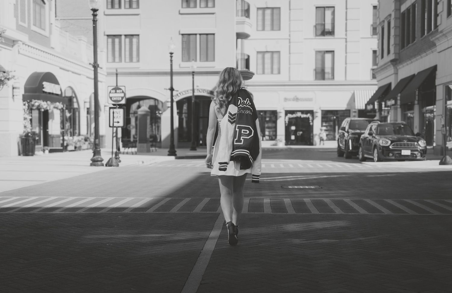 Black and white photo of a woman in a varsity jacket standing at a city street intersection.