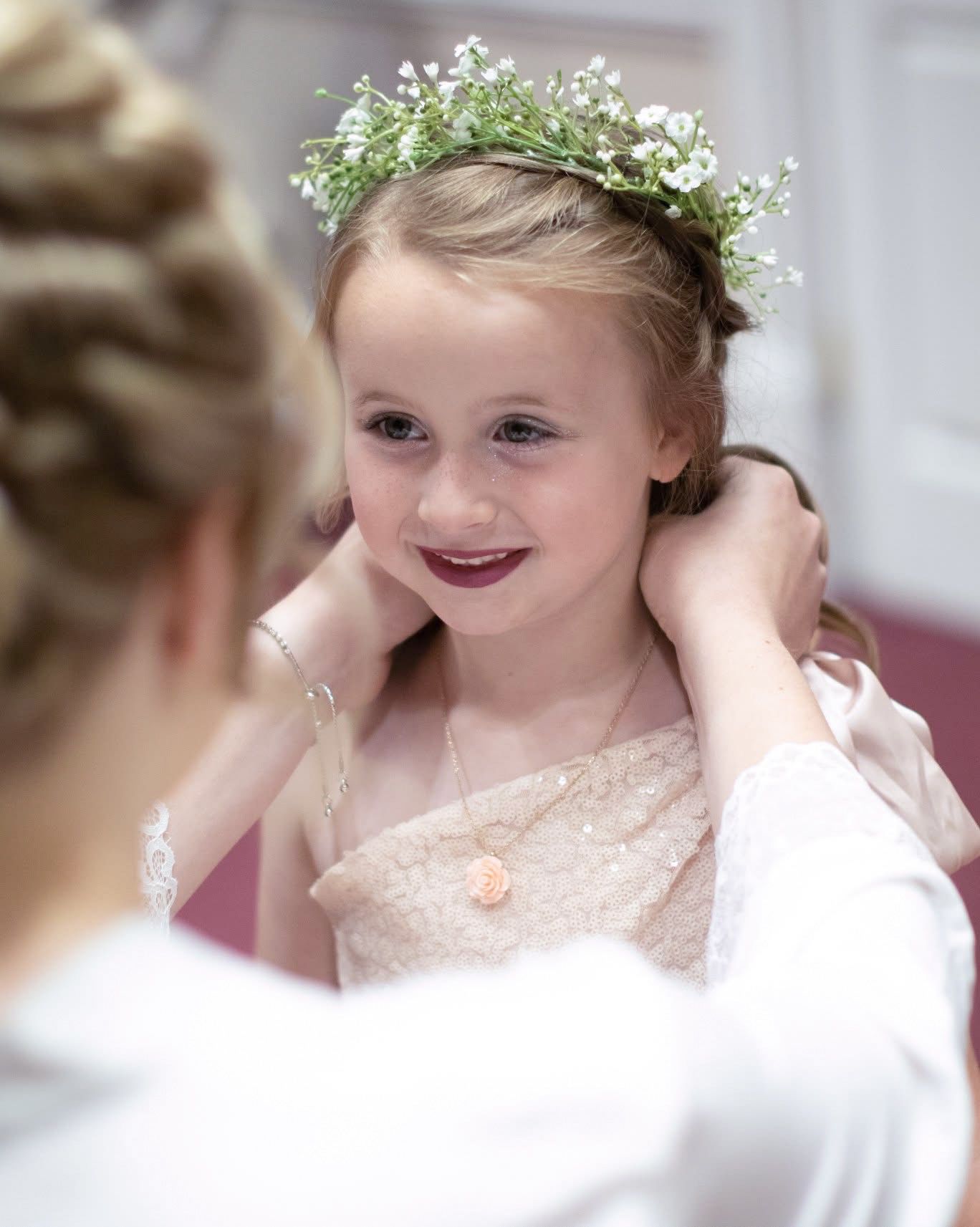 A young flower girl smiles as her hair is adjusted, wearing a delicate floral crown.