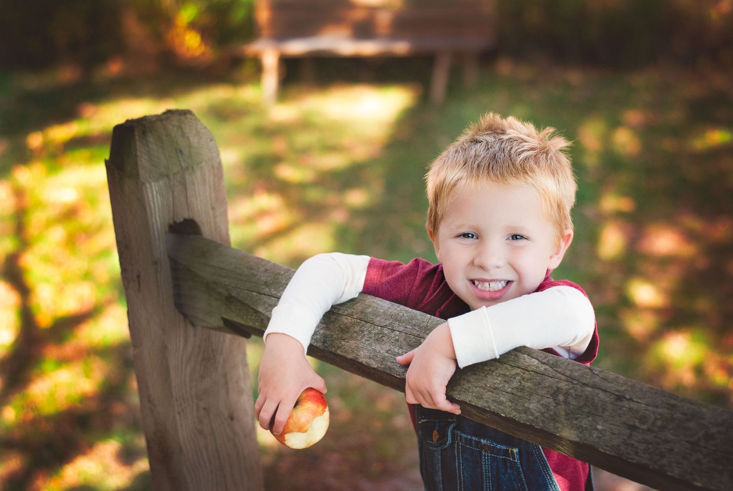 Happy blond boy smiling while leaning on wooden fence holding red apple outdoors.