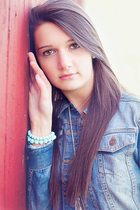 Beautiful teenage girl leaning against red wall in denim jacket senior portrait.