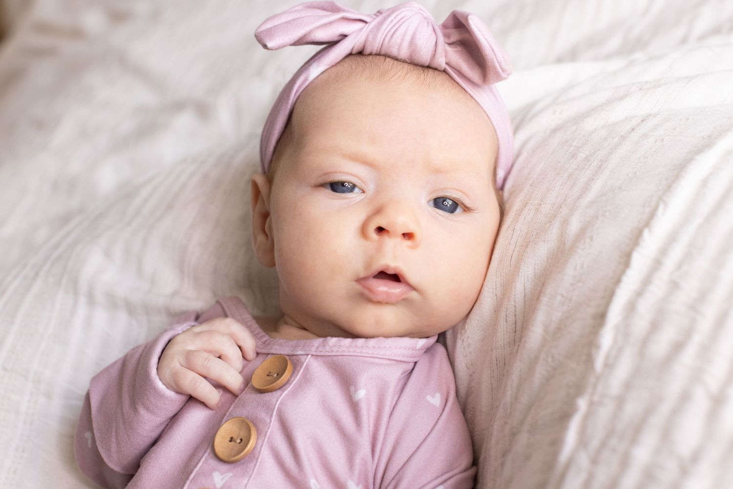 Newborn baby in pink outfit and bow headband lying on white blanket.
