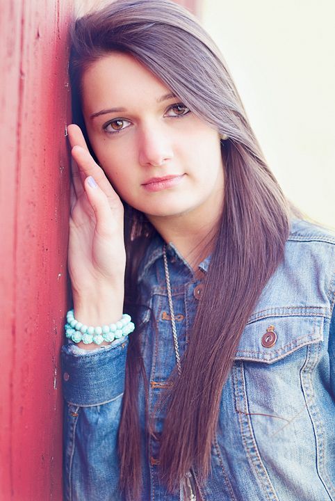 Young brunette woman in denim jacket leaning against red wall with hand on cheek.