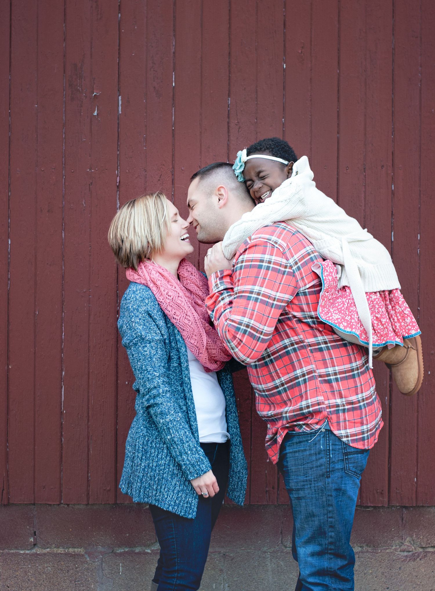 Happy interracial family with parents kissing laughing child against red barn wall.