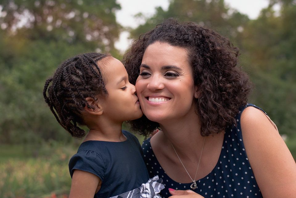 Young child kissing smiling mother on cheek outdoors in a green park setting.