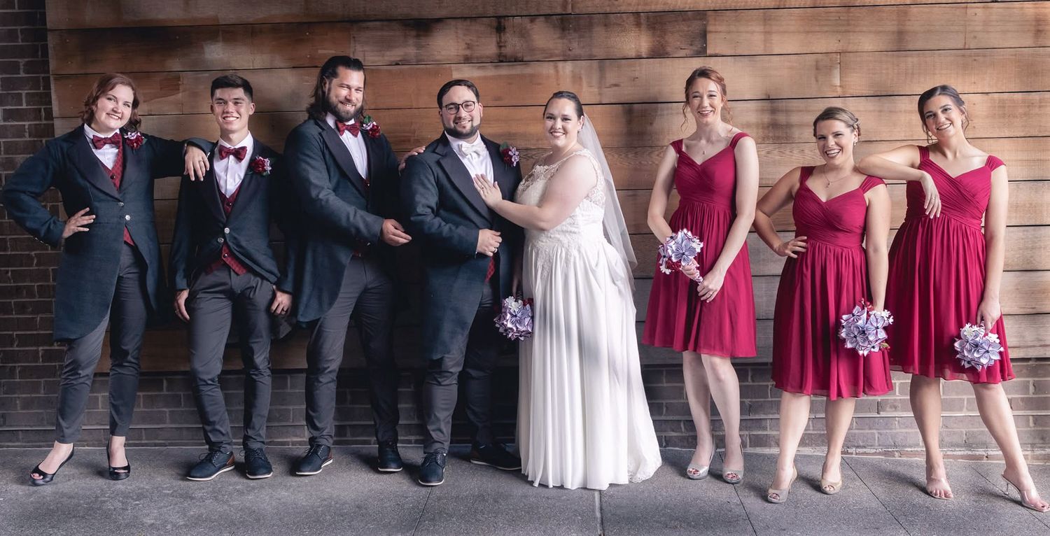Wedding party poses in burgundy and navy attire against rustic brick wall with bride in white gown.