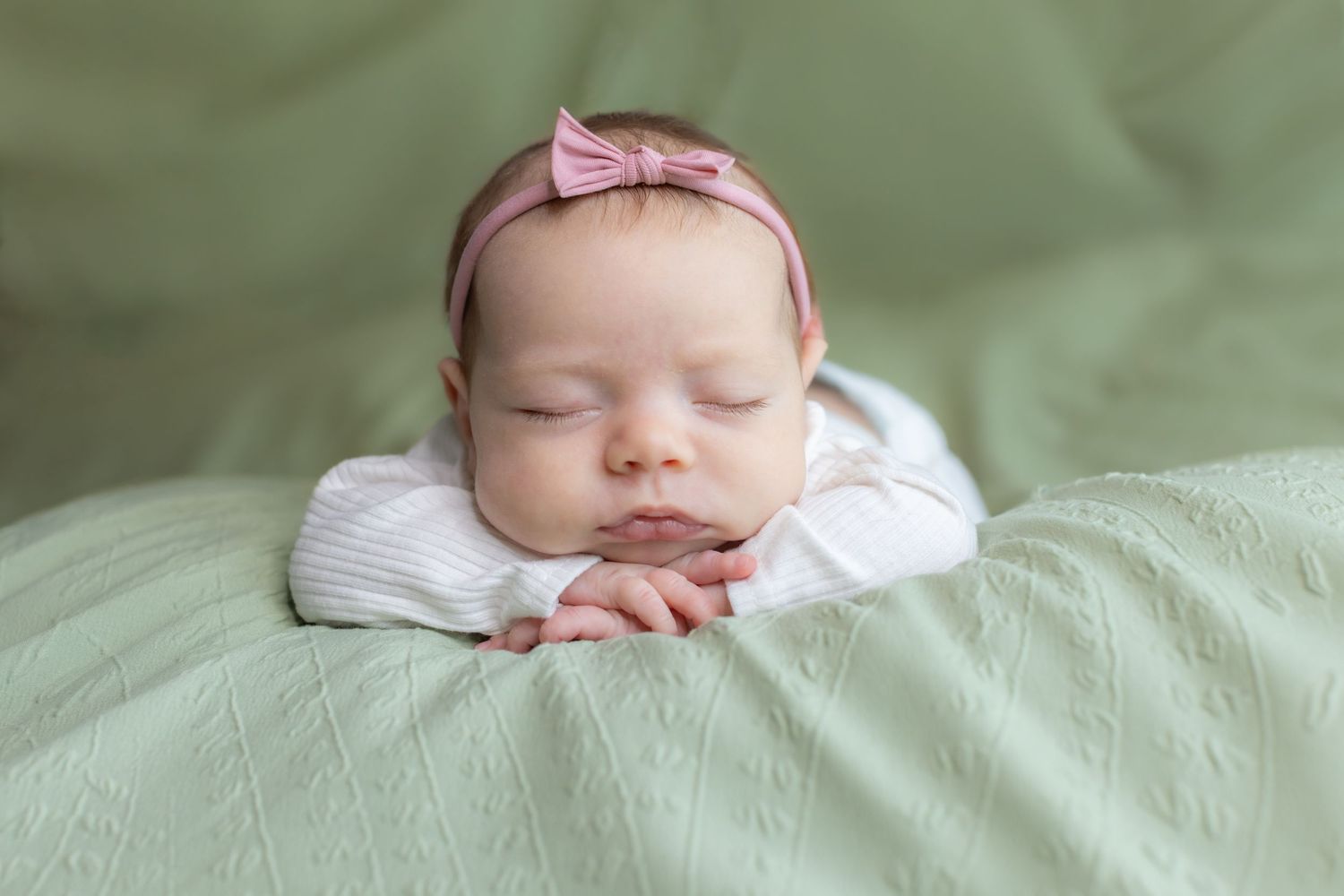 Peaceful newborn baby in white outfit sleeping on green blanket with pink bow headband.
