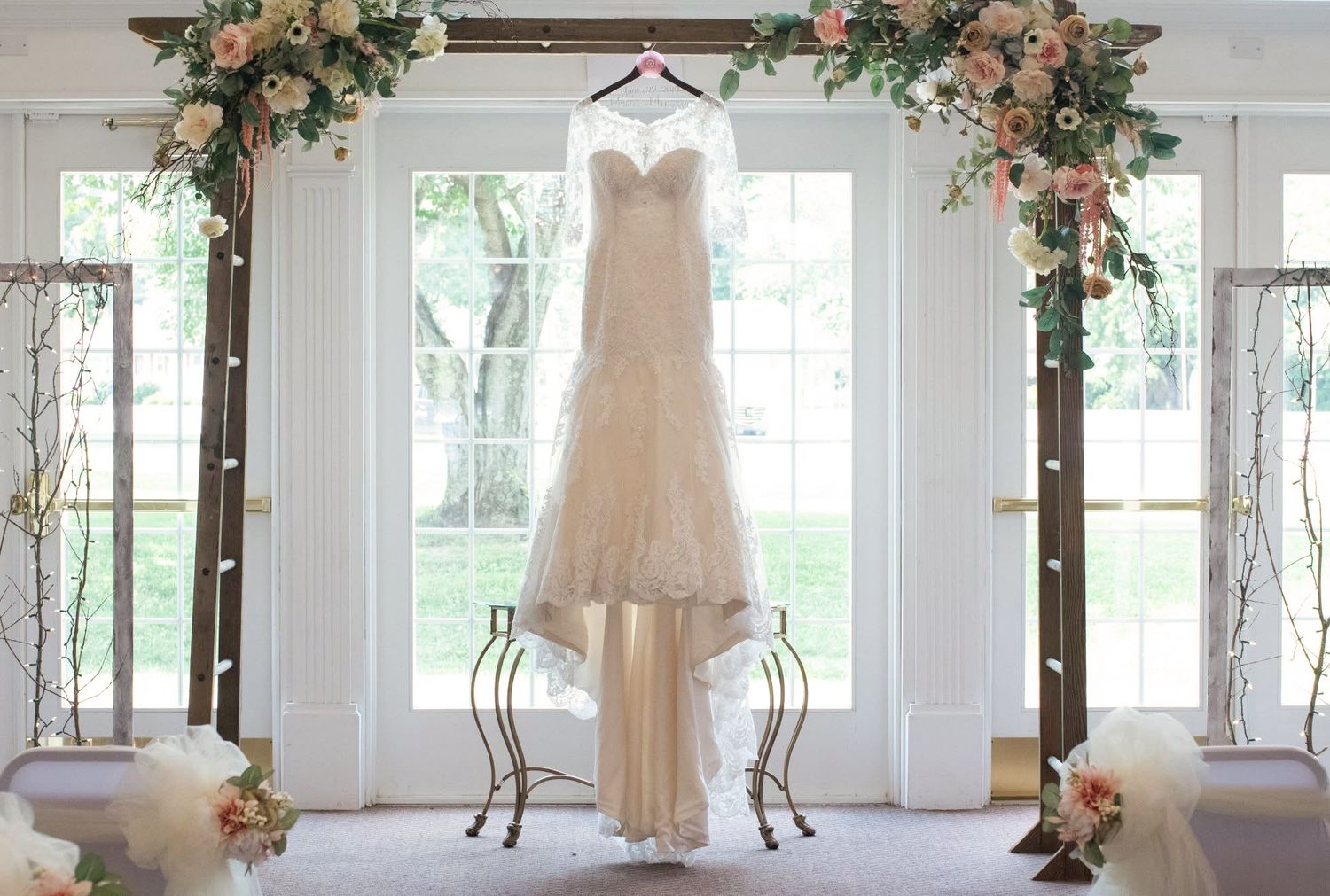 An elegant lace wedding dress displayed on a hanger in a floral-decorated ceremony venue.