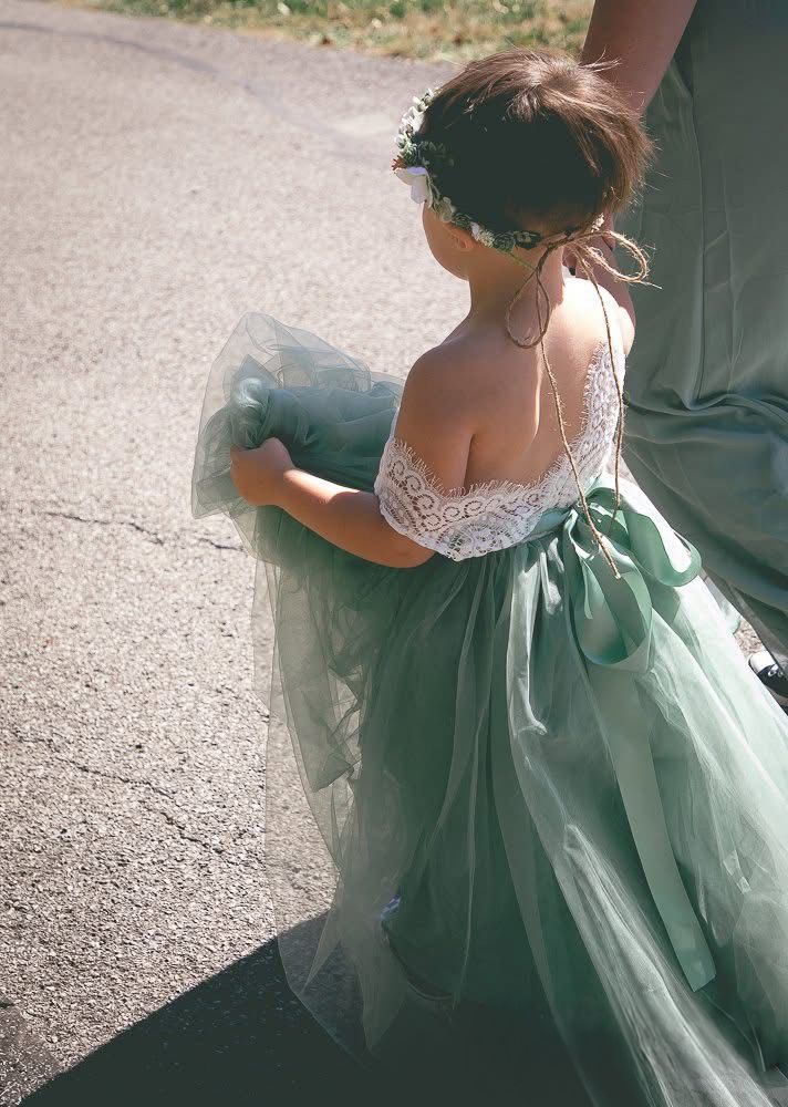 A young flower girl in a sage green tulle dress holds her skirt while walking outdoors.