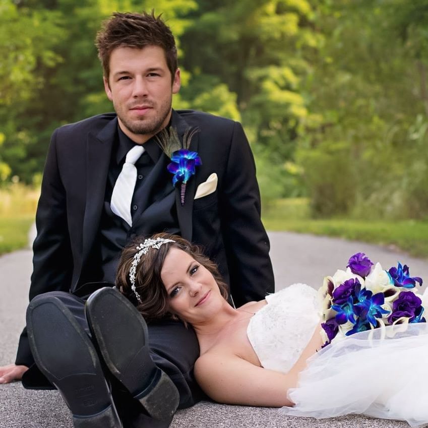Newlywed couple in wedding attire sitting on road surrounded by lush green trees.