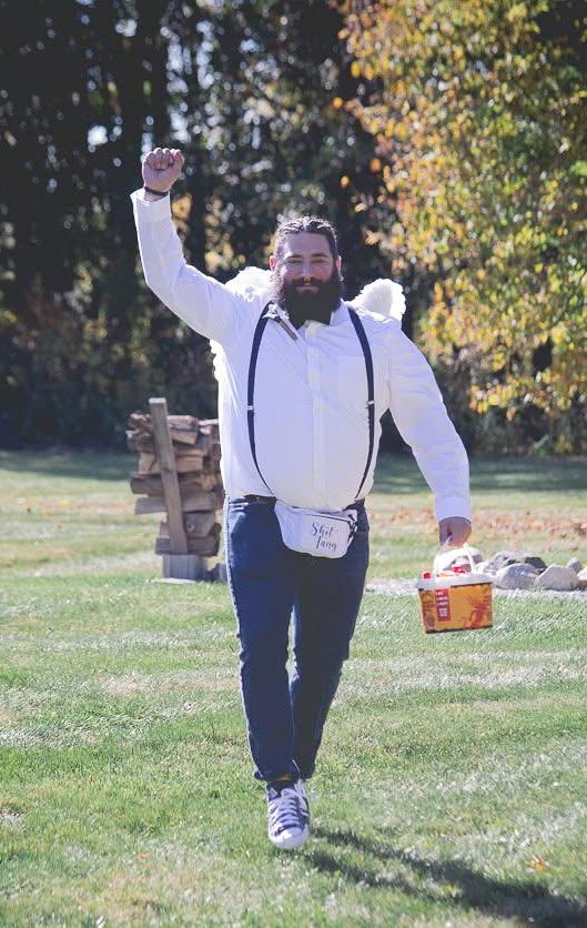 A bearded groomsman in suspenders cheers while carrying a basket on a sunny lawn.