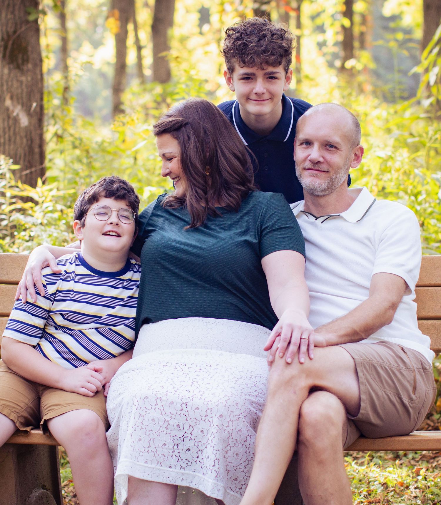 Happy family of four sitting together on bench surrounded by golden sunlit trees in park.