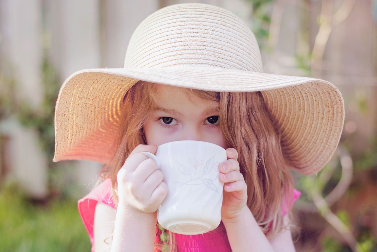 Young girl in wide brim straw hat drinking from white teacup in sunny outdoor garden setting.
