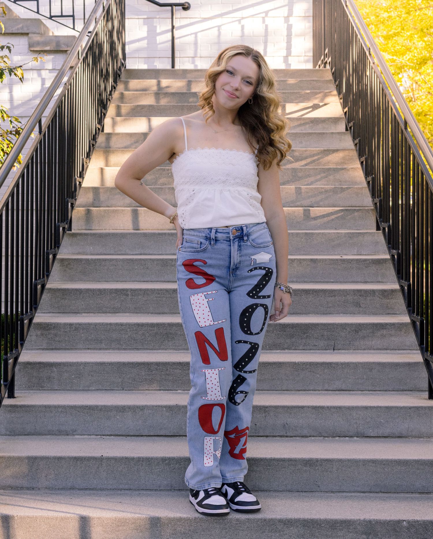 Young woman poses on stone steps wearing custom painted jeans reading SNOB 2026 with a white cami top.