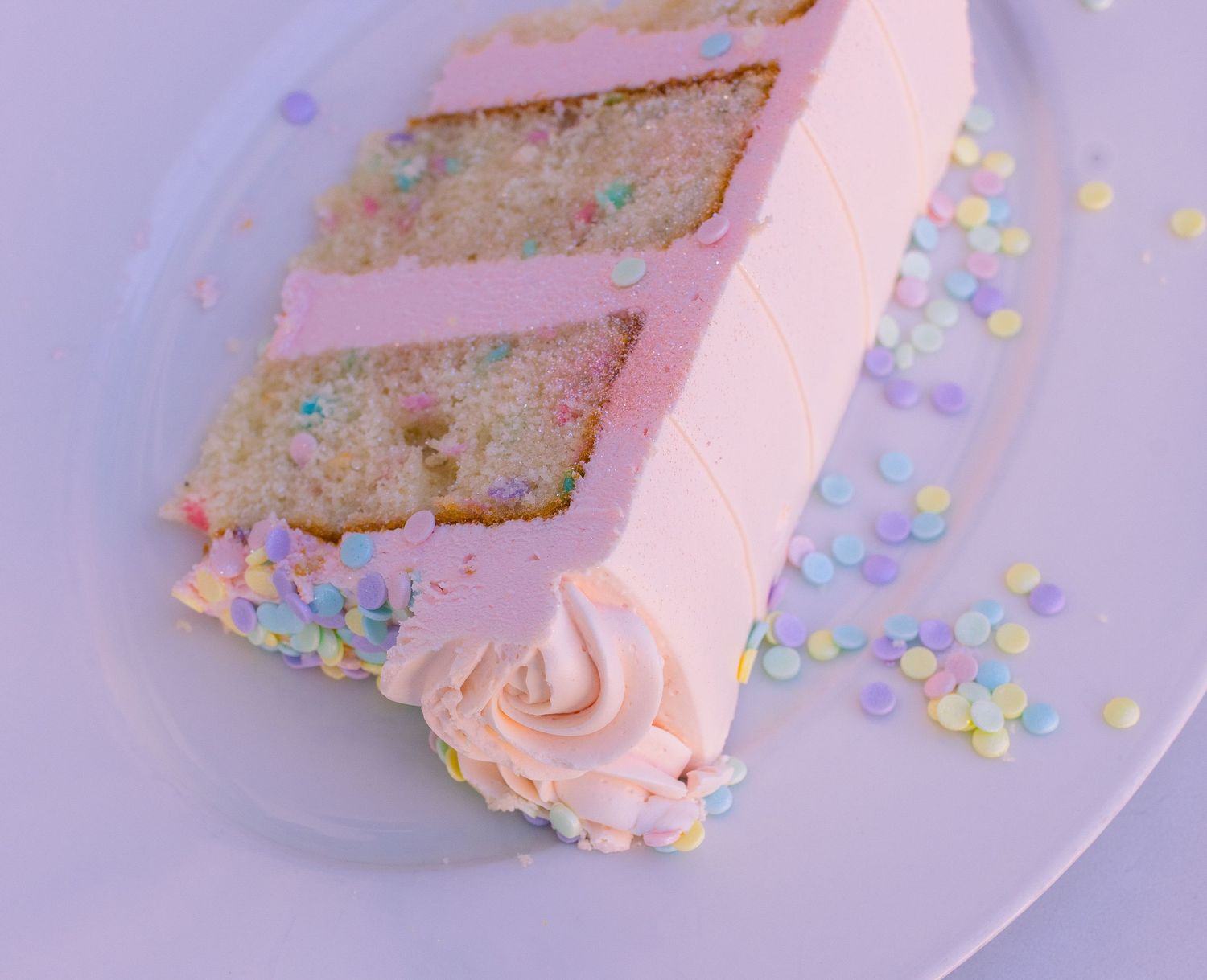 A slice of pastel pink layer cake with confetti sprinkles on a lavender plate.